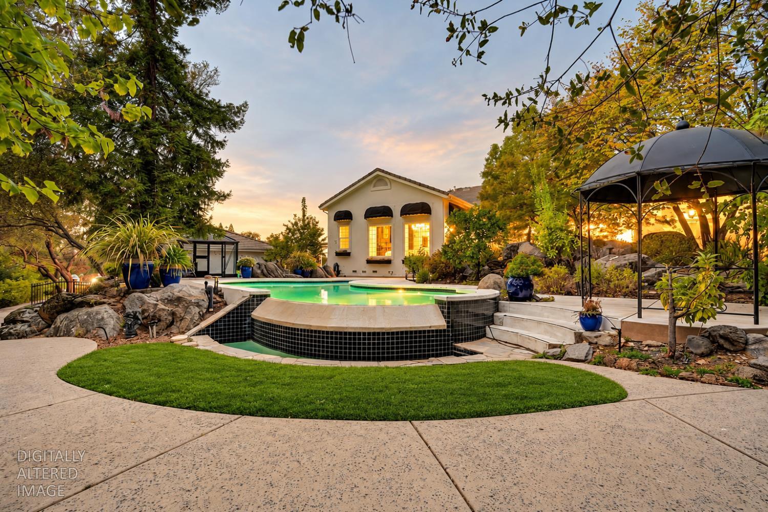 Backyard pool and raised spa with a black gazebo, stone landscaping, and fall-colored trees at sunset.