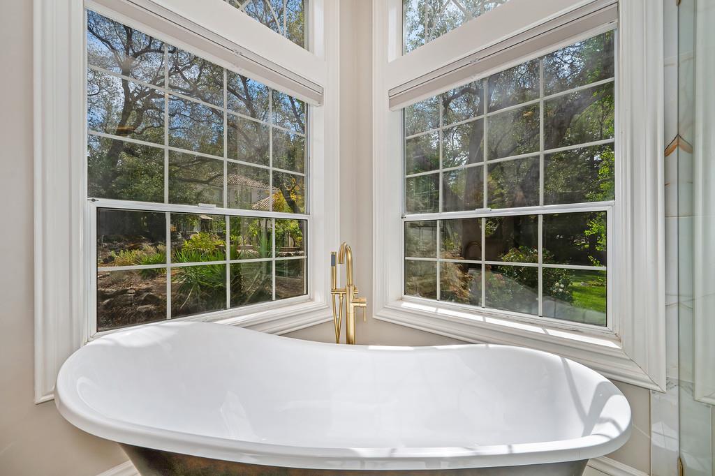 Freestanding soaking tub beneath corner windows overlooking a leafy garden.