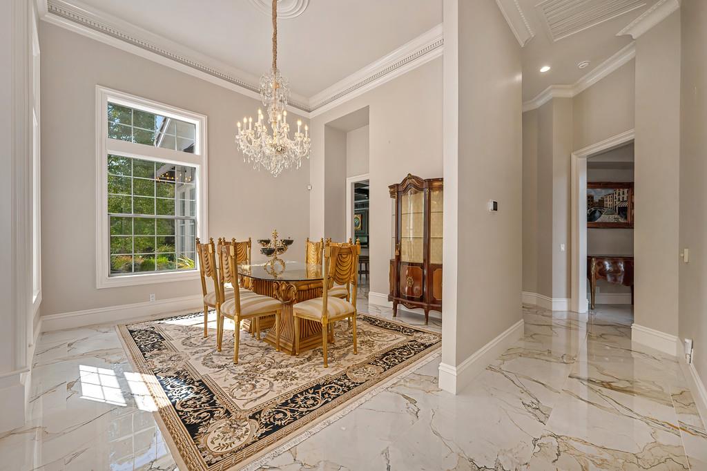 Formal dining room with a crystal chandelier, ornate rug, marble floors, and a large garden-facing window.