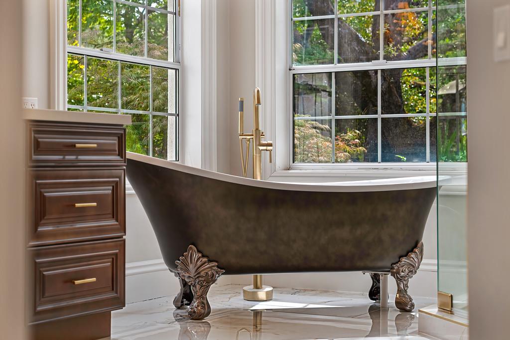 Bathroom with a dark clawfoot tub beneath large windows overlooking leafy trees.