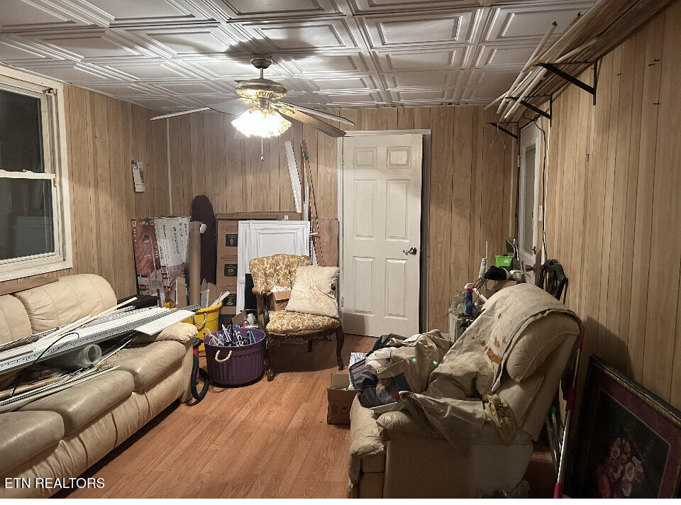 Cluttered living room with wood-paneled walls, a patterned ceiling, ceiling fan, and sofas.