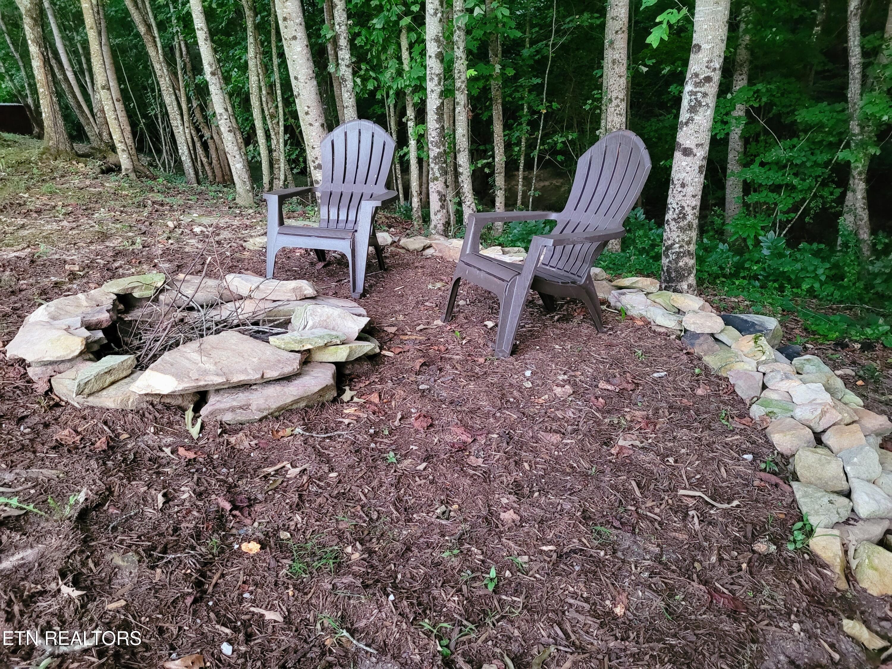 Two Adirondack chairs sit beside a stone-ringed fire pit in a wooded clearing.