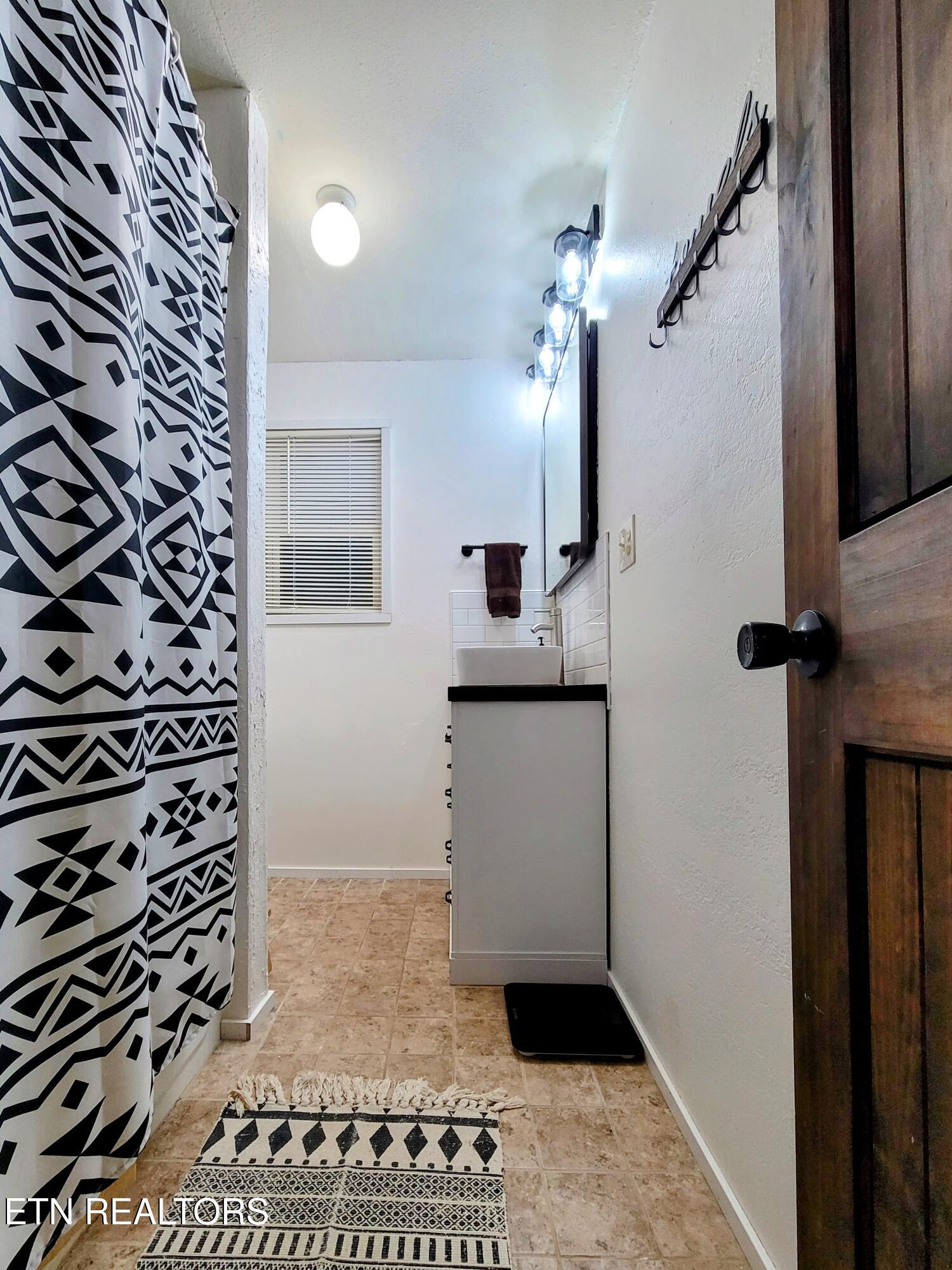 Bathroom with geometric shower curtain, white vessel sink, tiled floor, and patterned rugs.