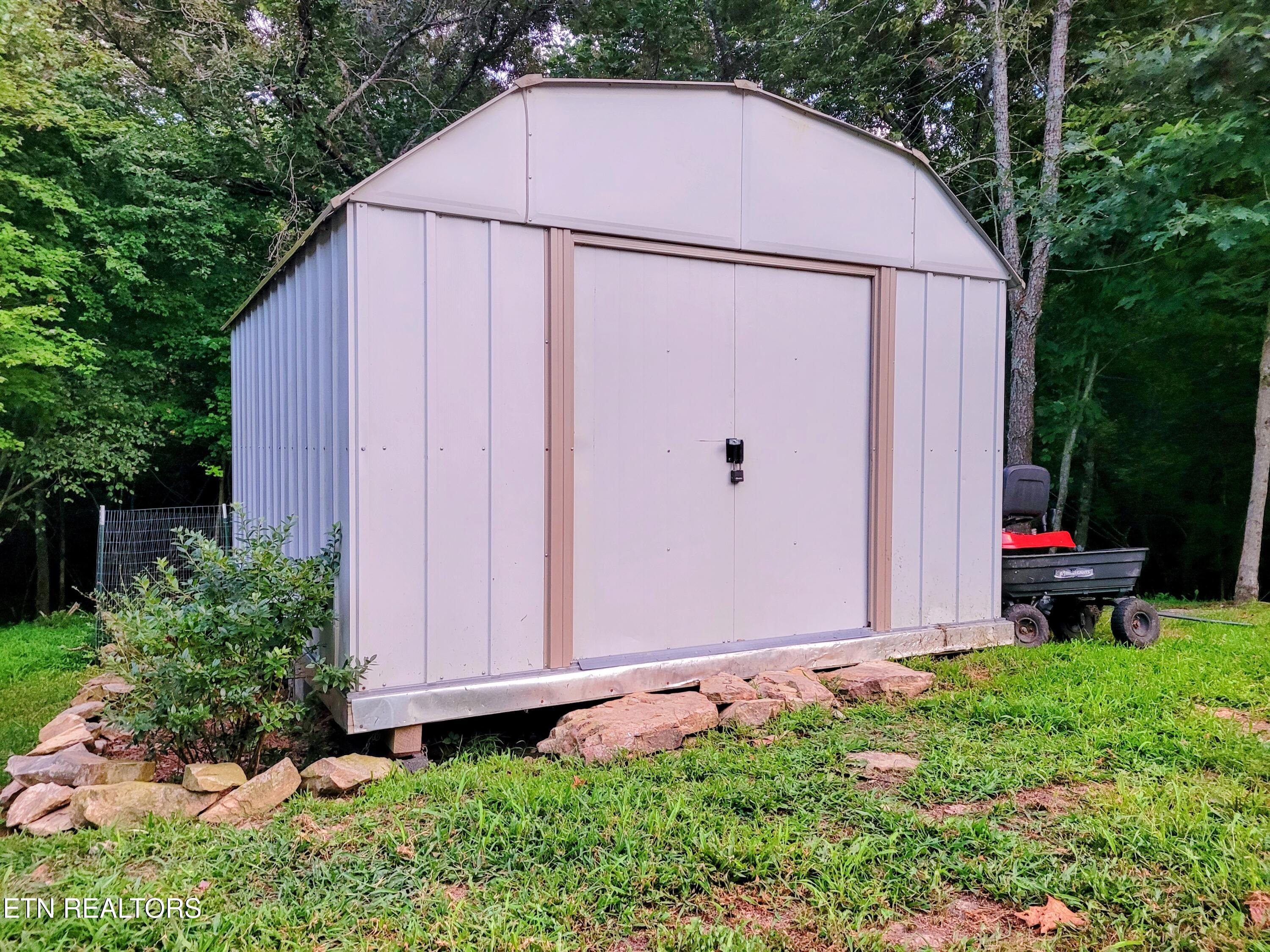 Metal storage shed with double sliding doors on a grassy, wooded lot, with rocks along its base.