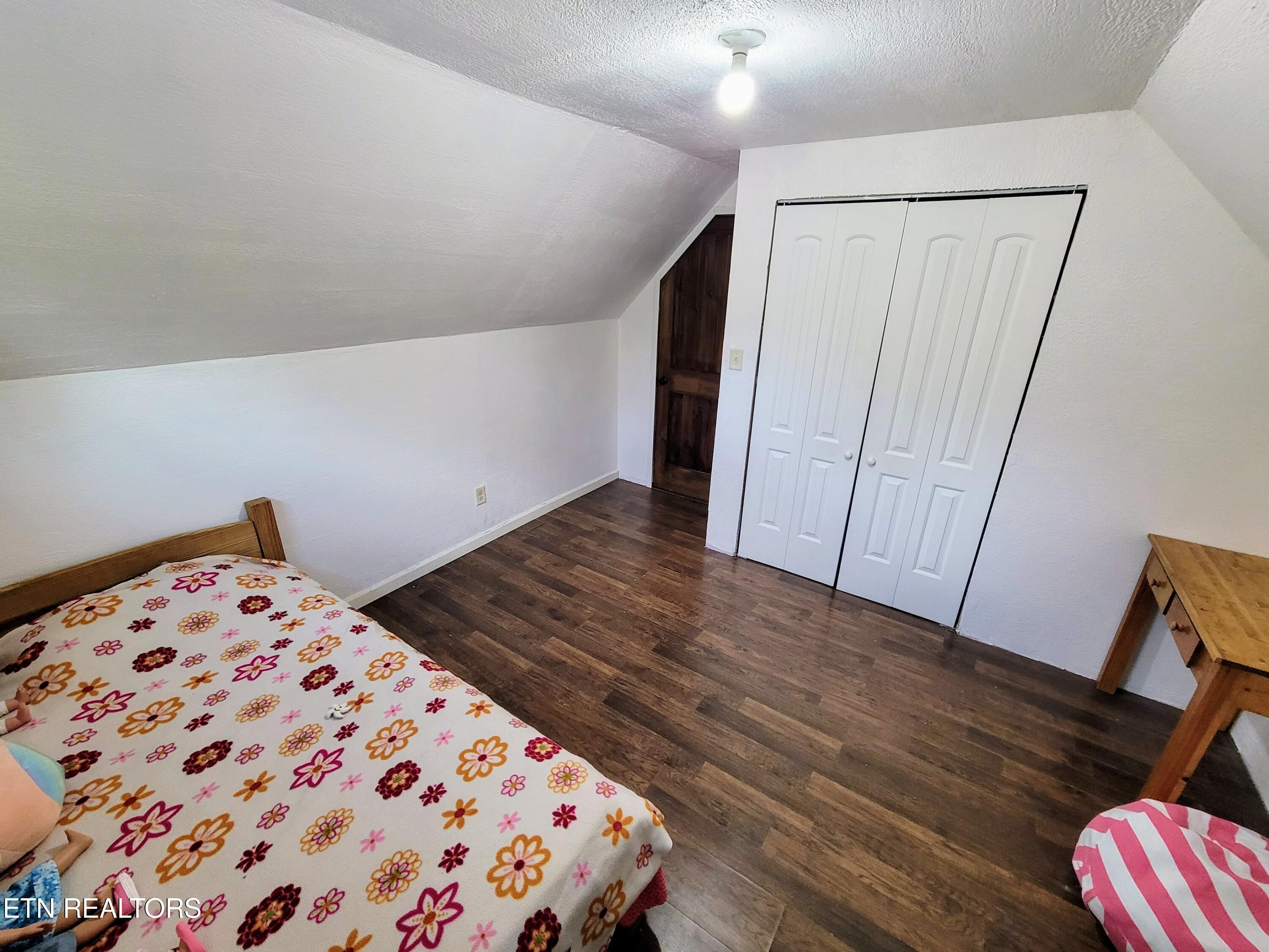 Attic bedroom with sloped ceiling, floral bedding, white closet doors, and dark wood-look flooring.