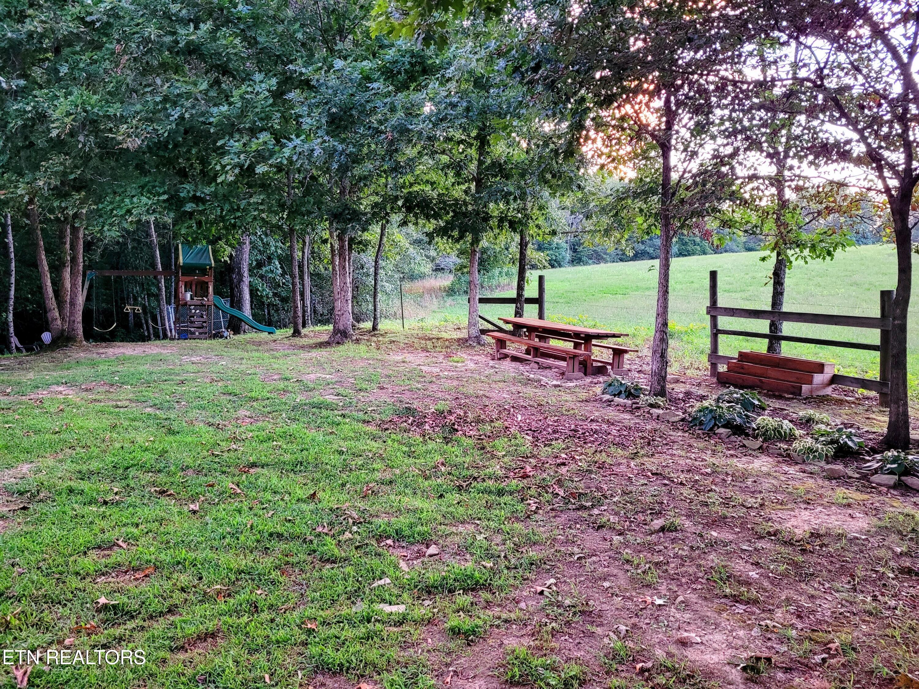 Tree-shaded yard with a picnic table, raised garden bed, and children’s playset beside an open field.