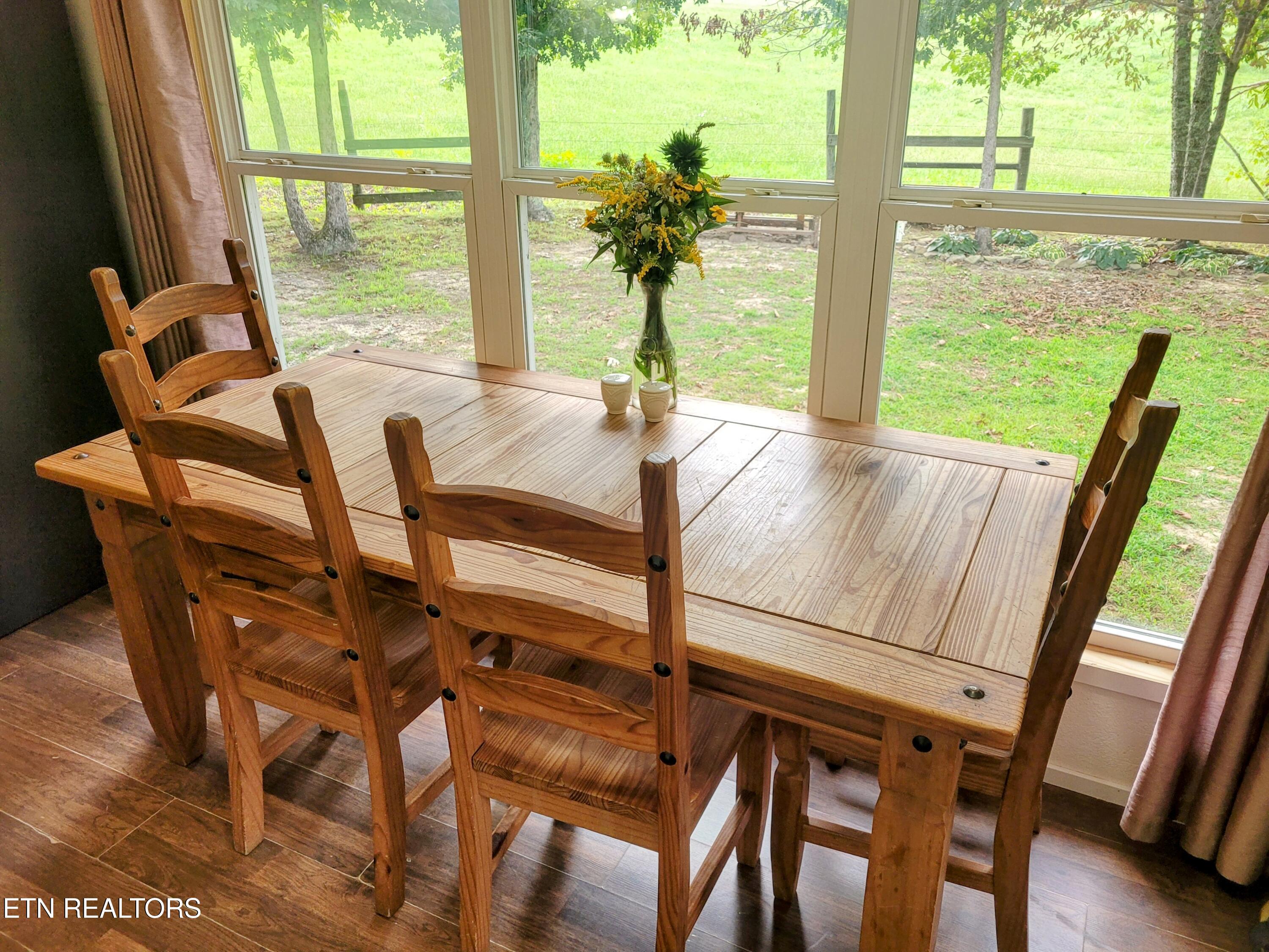 Dining room with a rustic wooden table and chairs beside large windows overlooking a green yard.