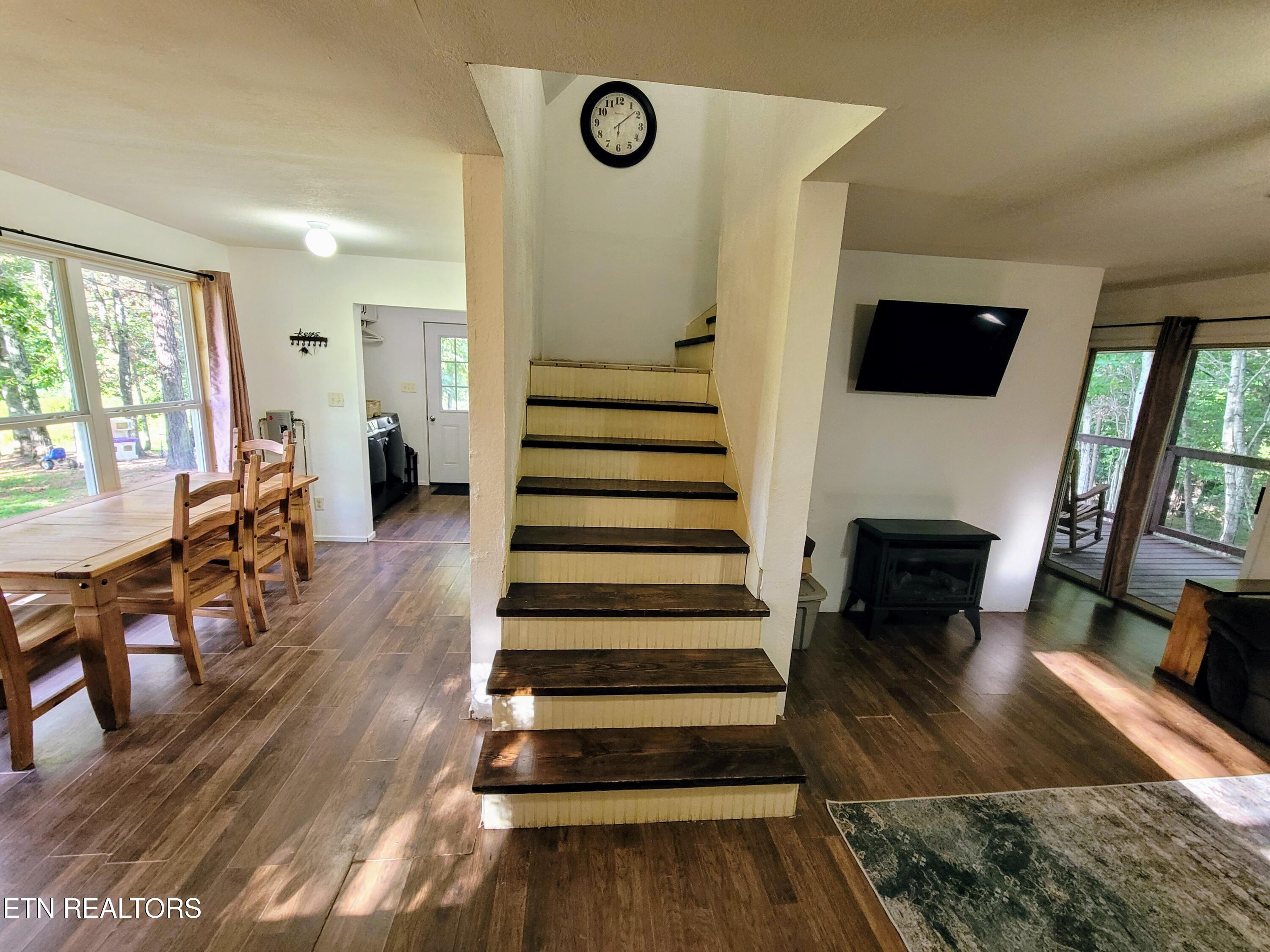 Open living and dining area with a central staircase, wood flooring, and windows overlooking trees.