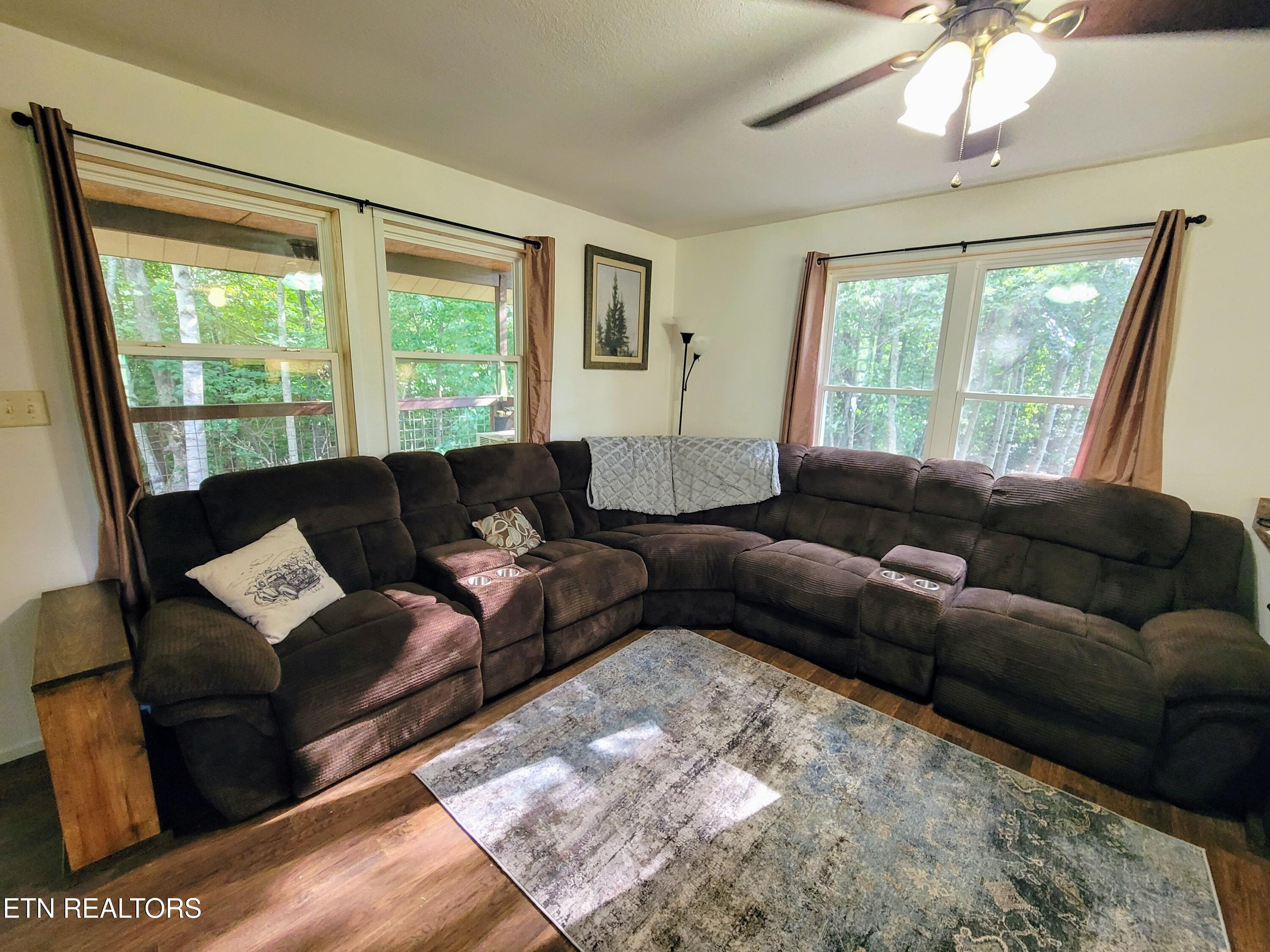 Living room with a dark sectional sofa, patterned rug, and windows overlooking wooded surroundings.