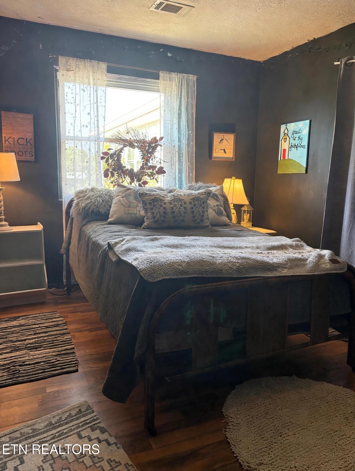 Furnished bedroom with a dark bed, layered gray bedding, and a bright window with sheer curtains.