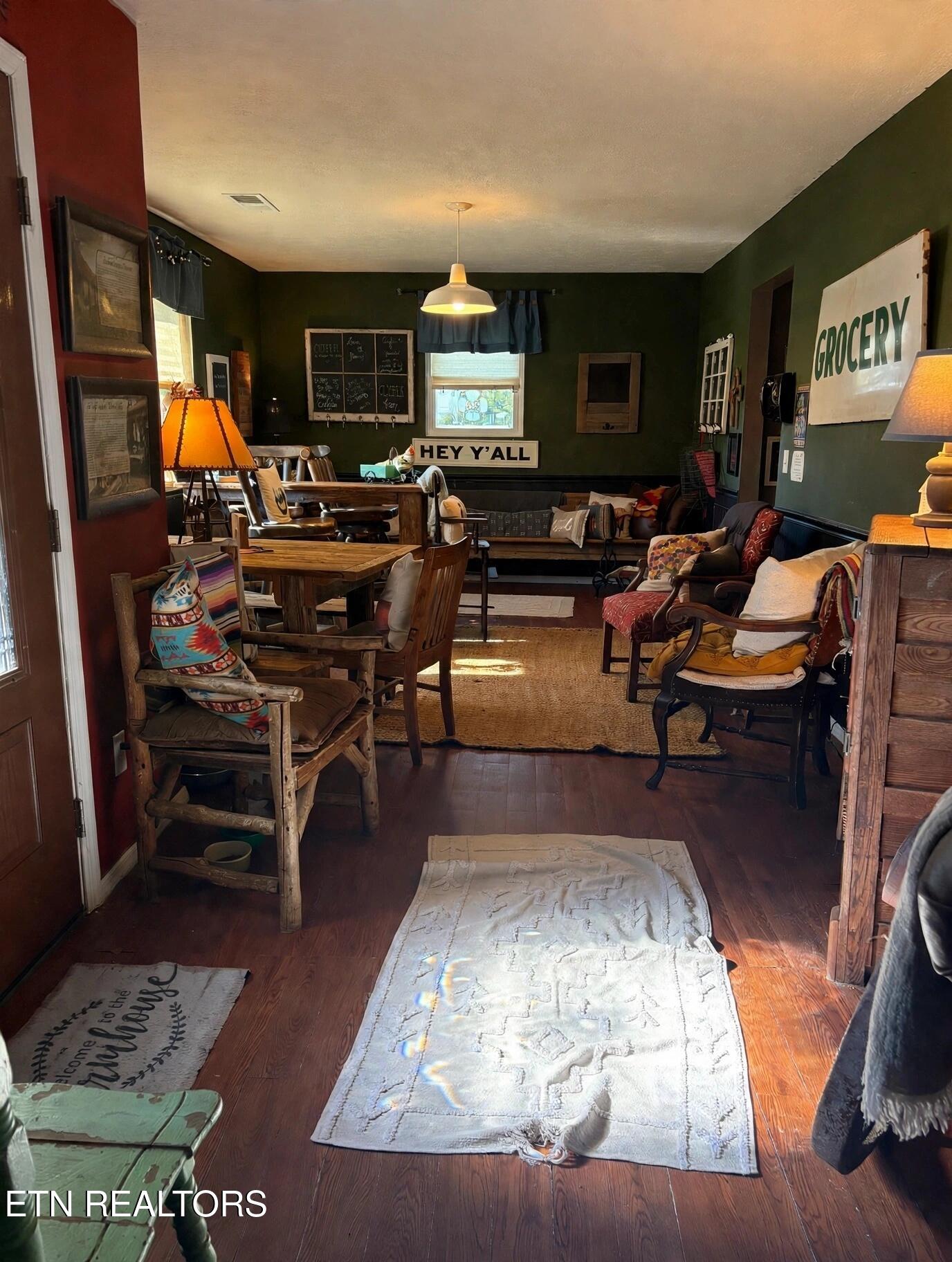 Furnished living room with dark green walls, wood floors, colorful cushions, and a “HEY Y’ALL” sign.