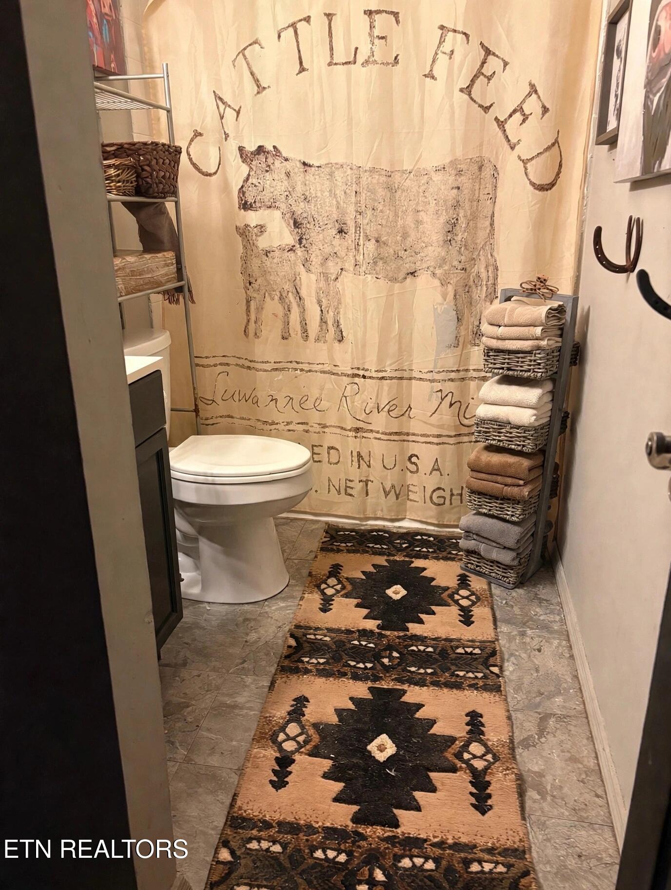 Small bathroom with a toilet, cattle feed sack shower curtain, and patterned runner.