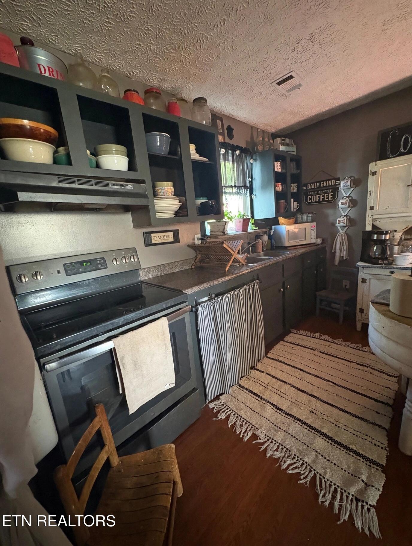 Kitchen with dark open shelving, stainless steel range, sink beneath a curtained window, and striped runner rug.