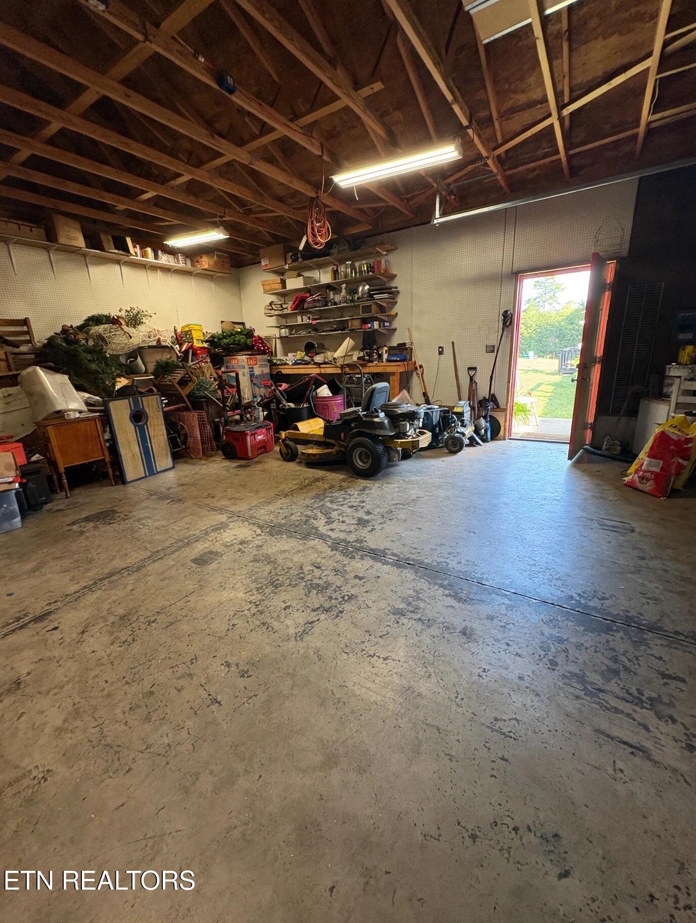 Garage workshop with a riding mower, storage shelves, exposed rafters, and an open doorway to the yard.