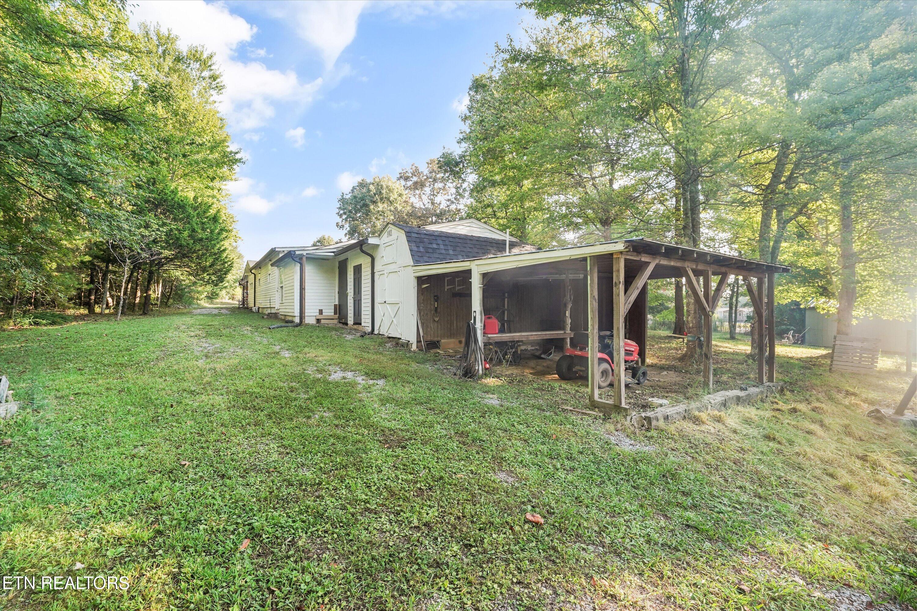 House beside a large wooden carport, with a red riding mower and mature trees around the grassy yard.