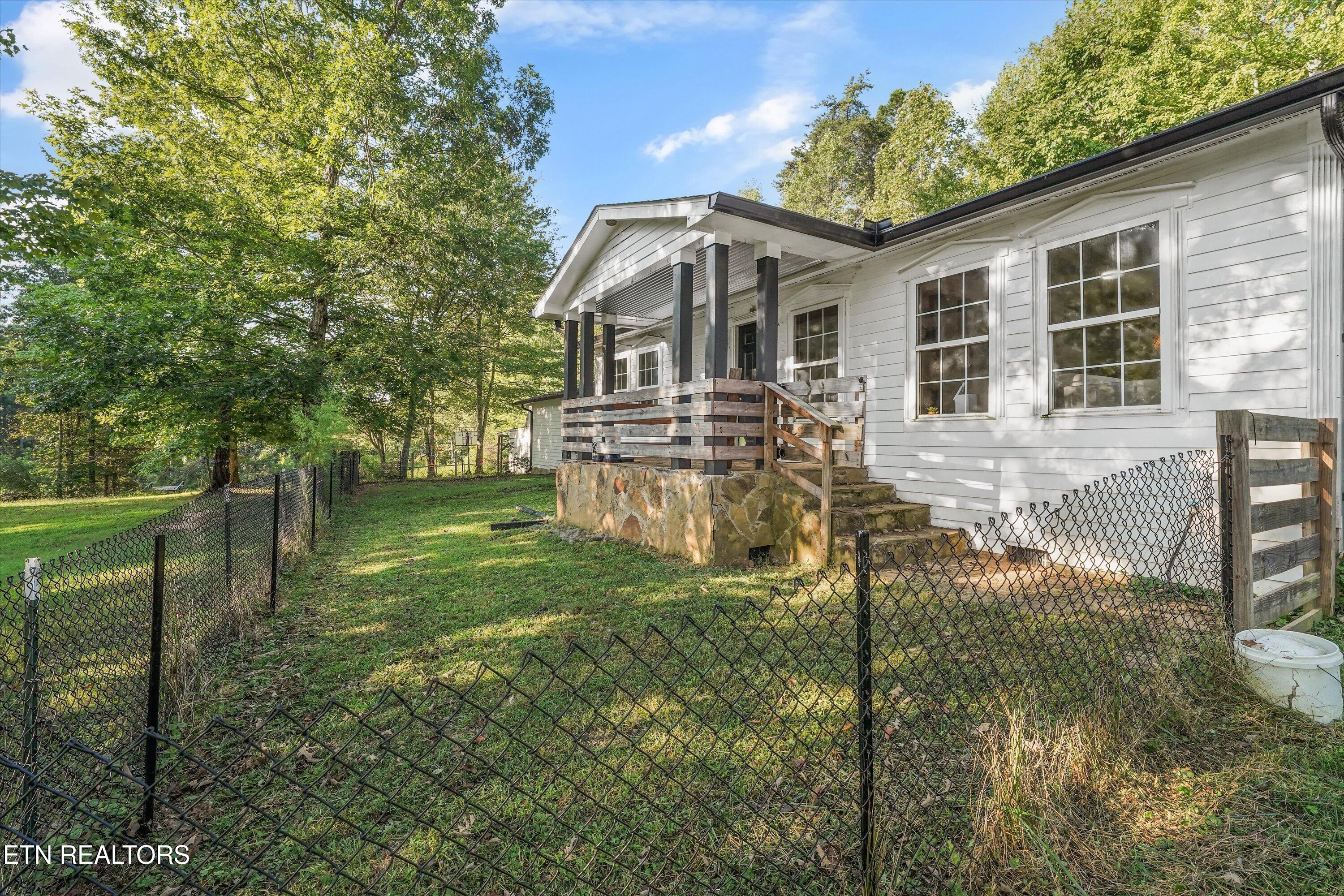 White single-story house with a covered wooden porch behind a chain-link fence and leafy trees.