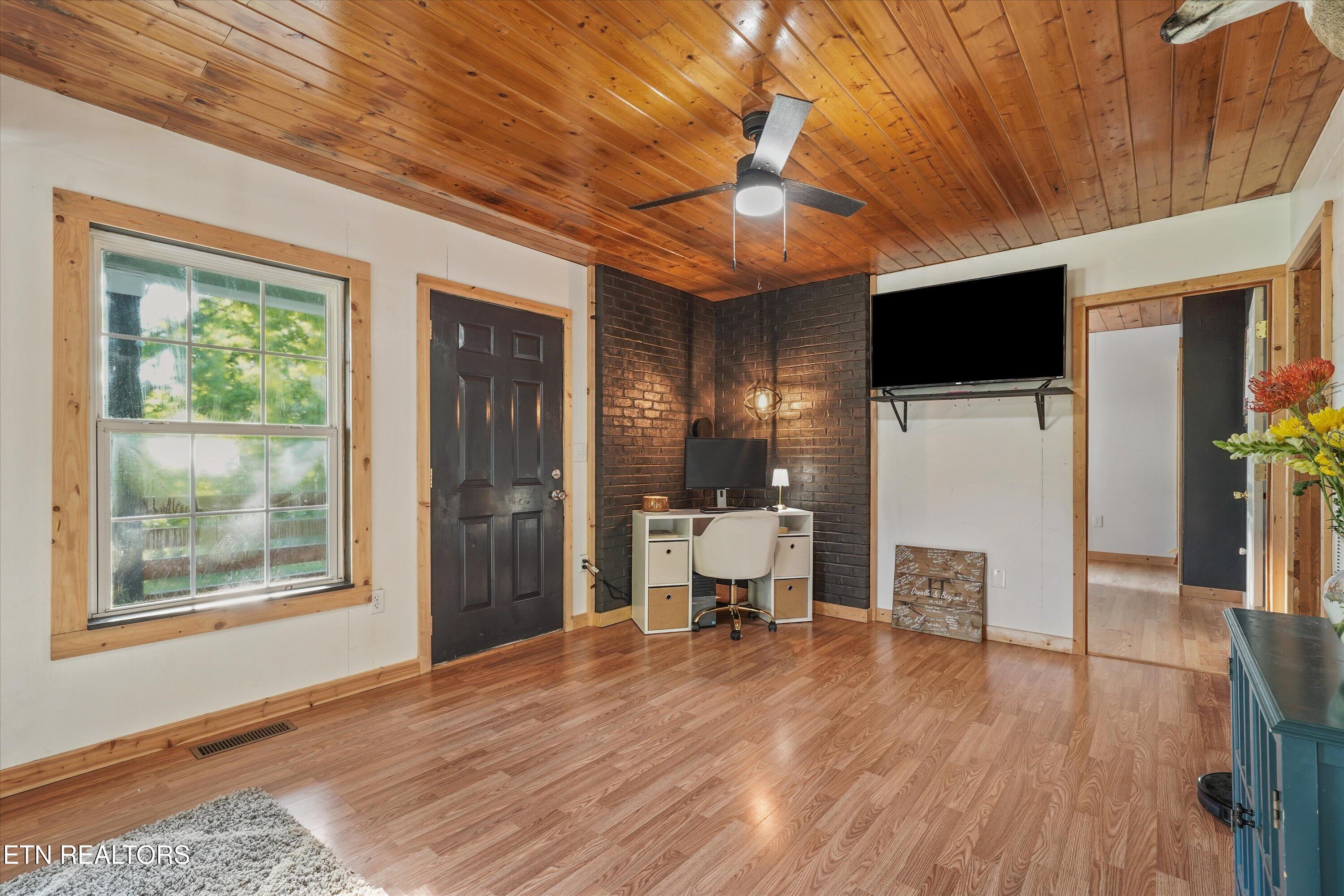Furnished room with a desk, dark brick accent wall, wall-mounted television, and wood-plank ceiling.