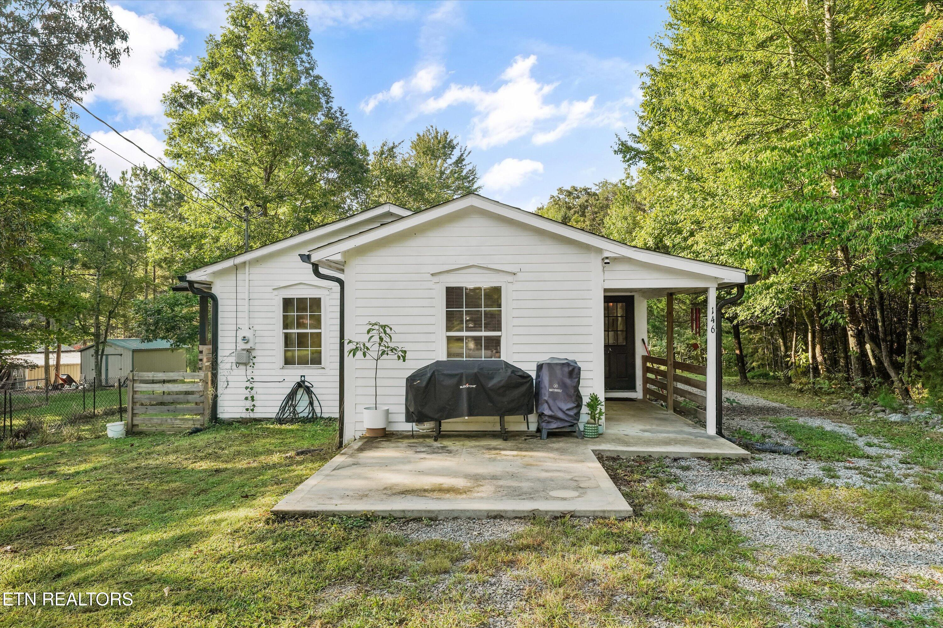 White single-story house with a covered entry, two covered grills, and a concrete driveway among trees.