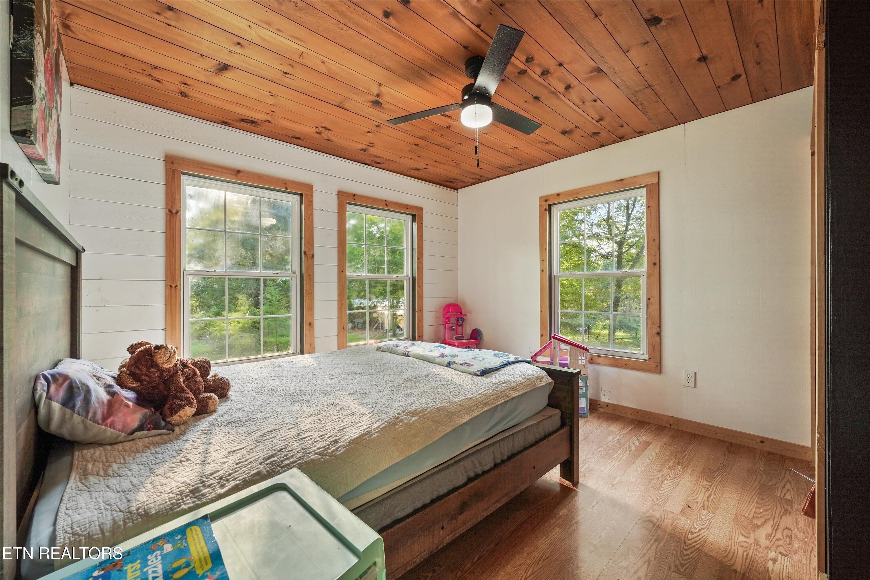 Bedroom with a wood-plank ceiling, three windows, and a large bed with a teddy bear.