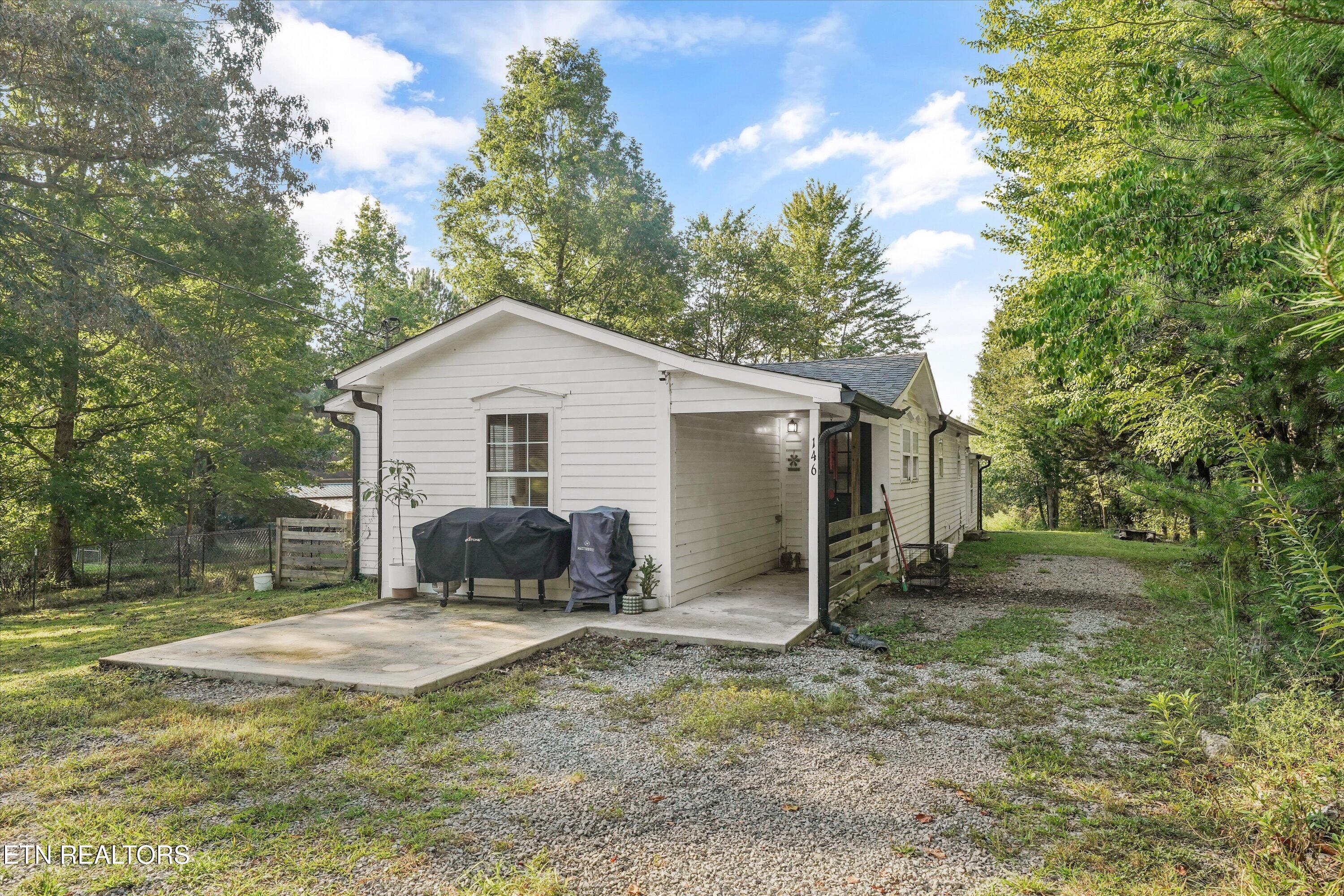 White single-story house with a covered carport, concrete patio, and gravel driveway among mature trees.