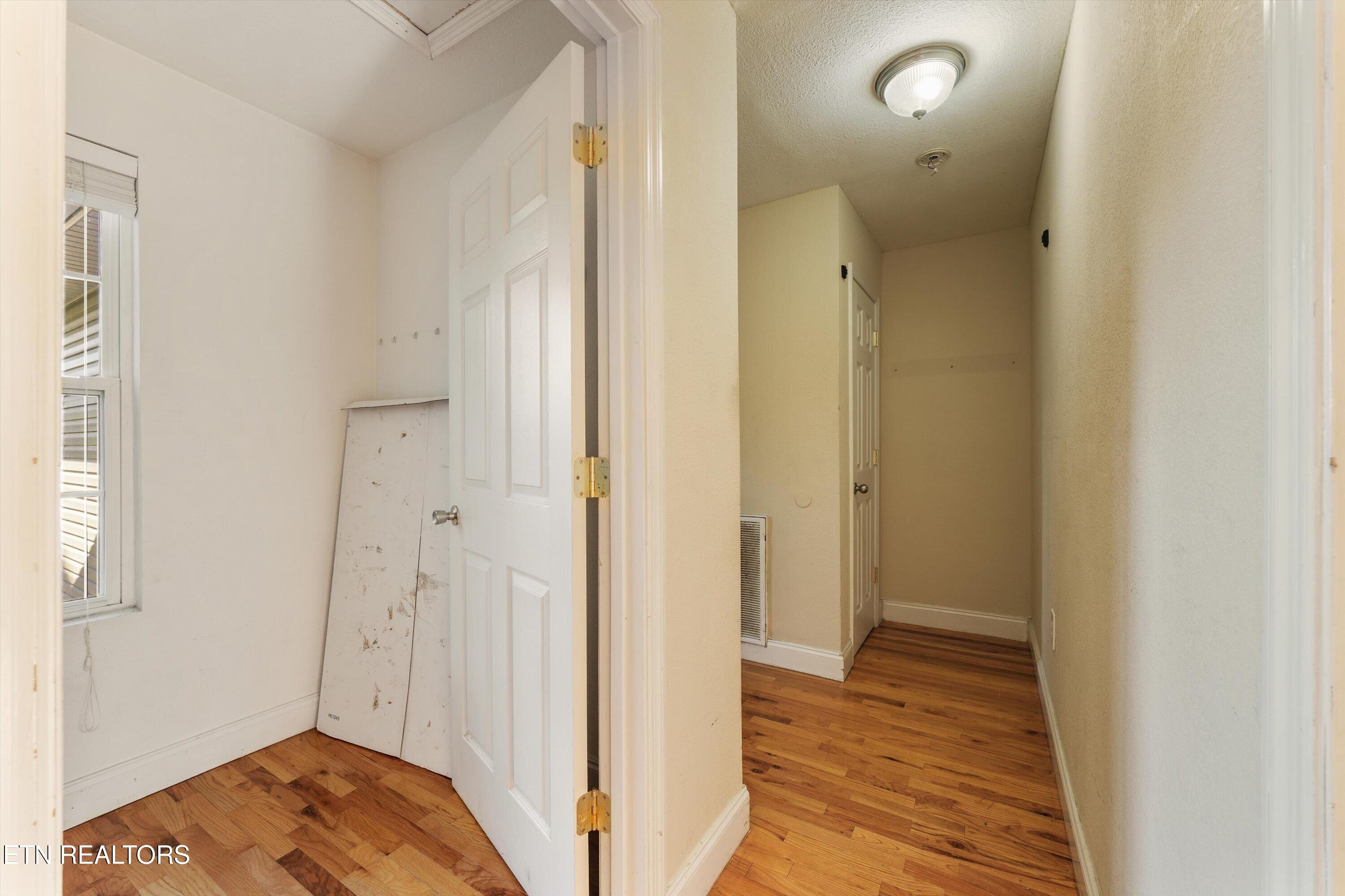Vacant hallway and adjoining room with hardwood floors, white doors, and a window with blinds.