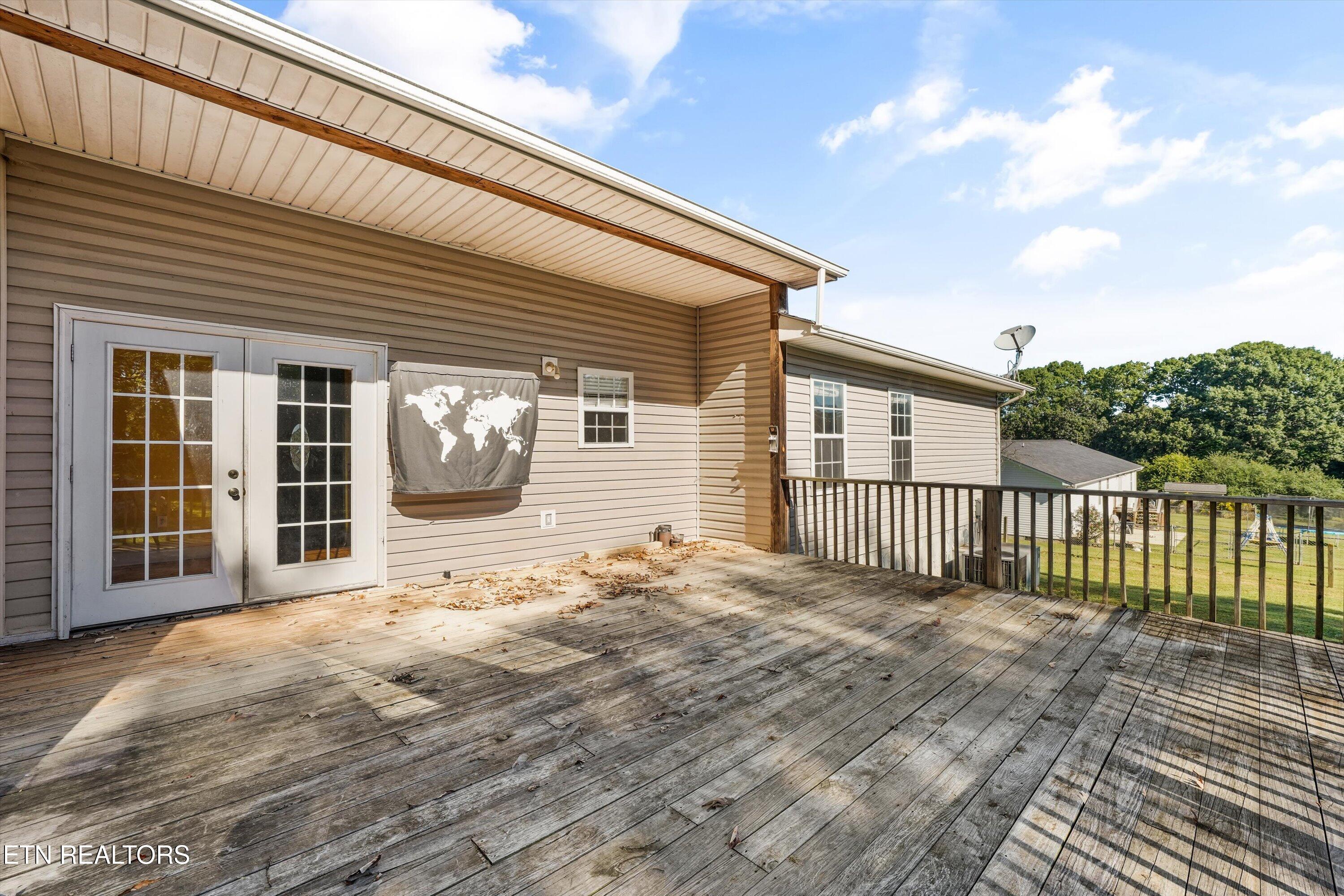 Weathered wooden deck along a beige-sided home, covered by a broad roof and overlooking a green yard.
