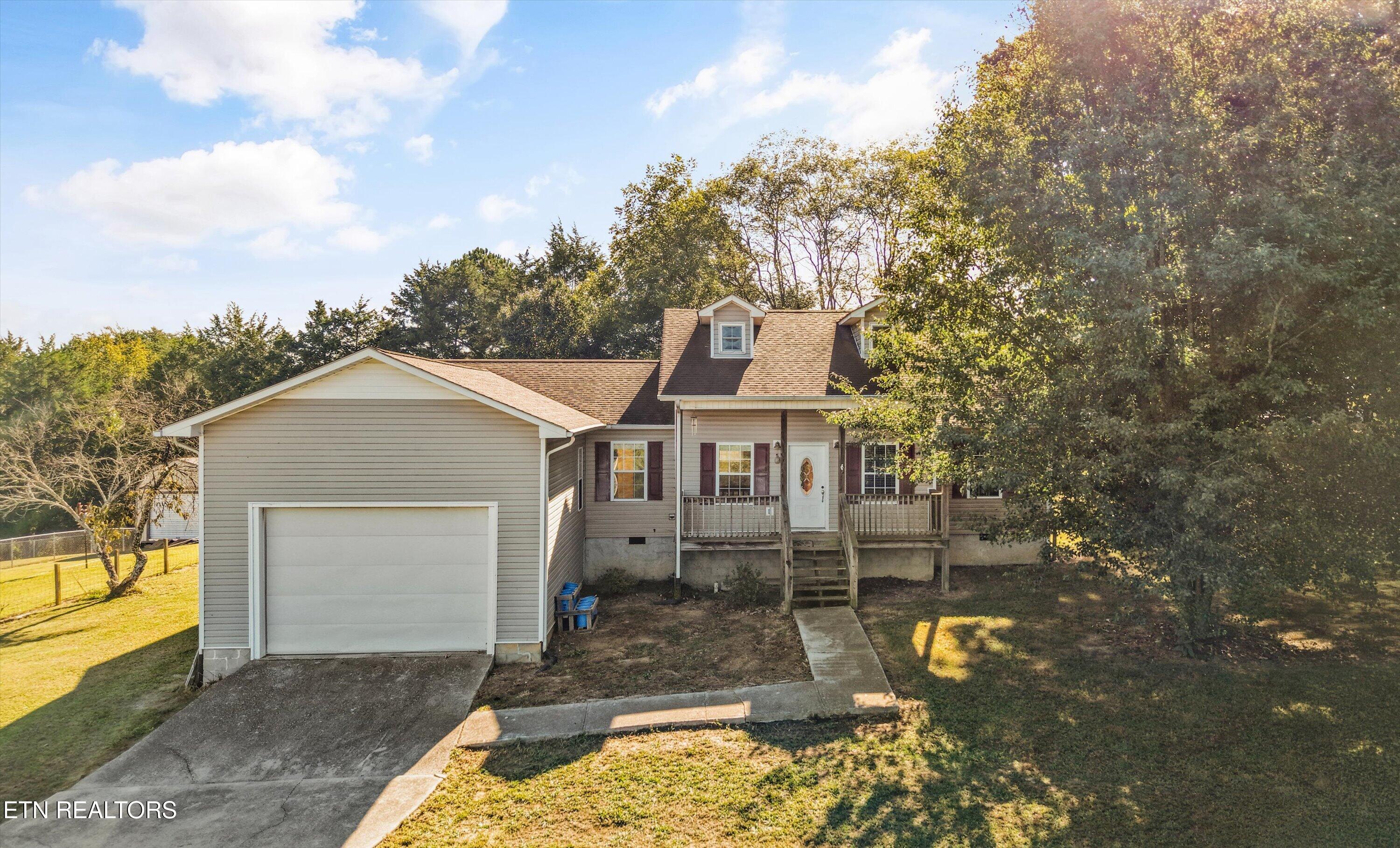 Gray-sided house with attached garage and covered porch beneath a large leafy tree.
