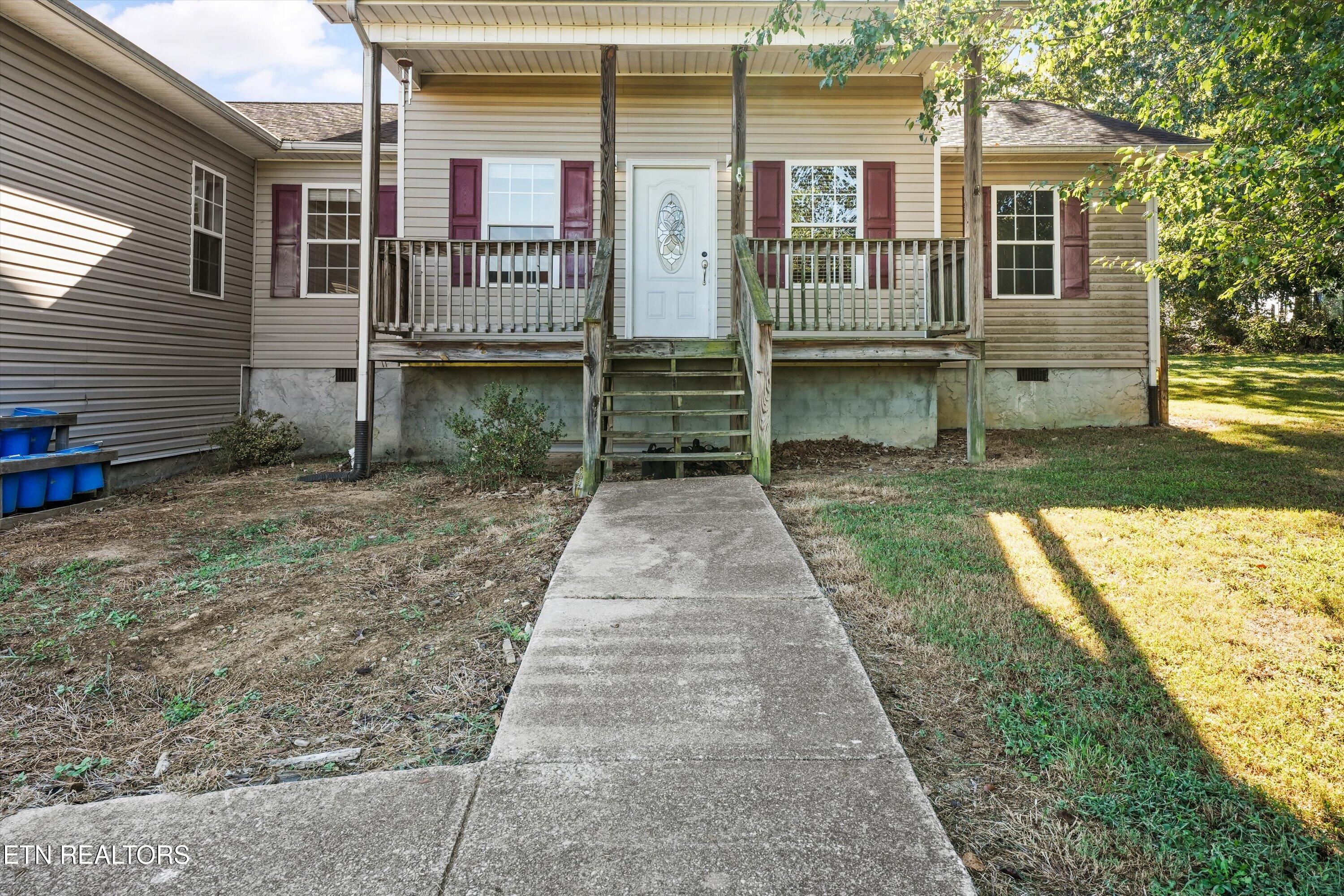 Beige-sided house with burgundy shutters and a raised covered porch above a concrete walkway.