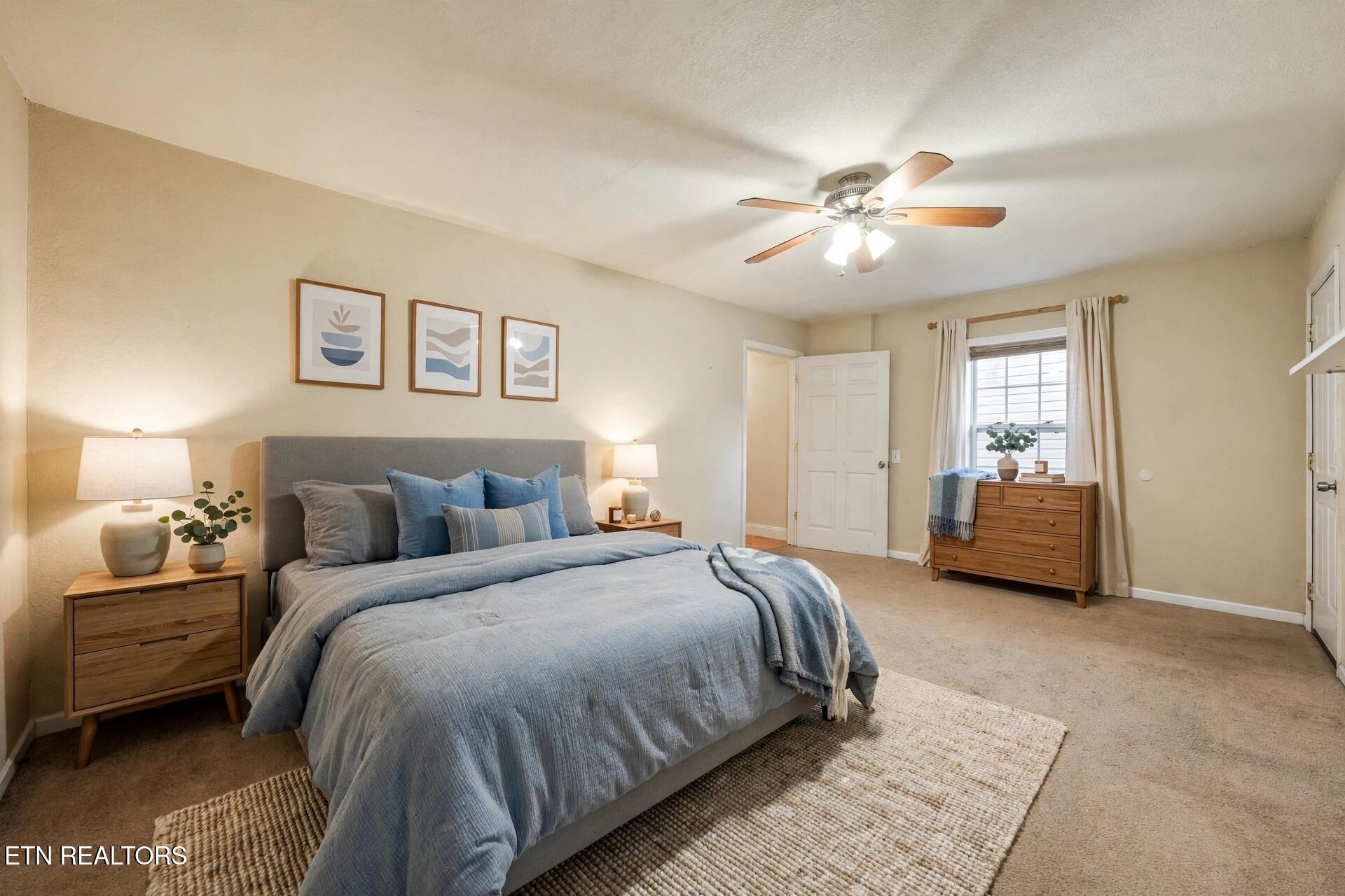 Staged bedroom with blue bedding, wood furniture, and a curtained window beside the dresser.