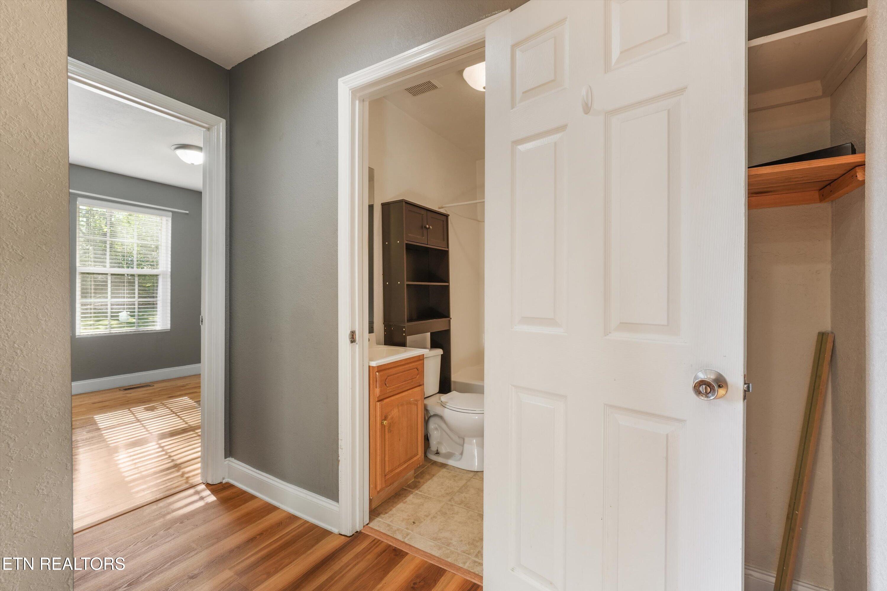 Bathroom doorway with a wood vanity and toilet beside a sunlit room with hardwood floors.