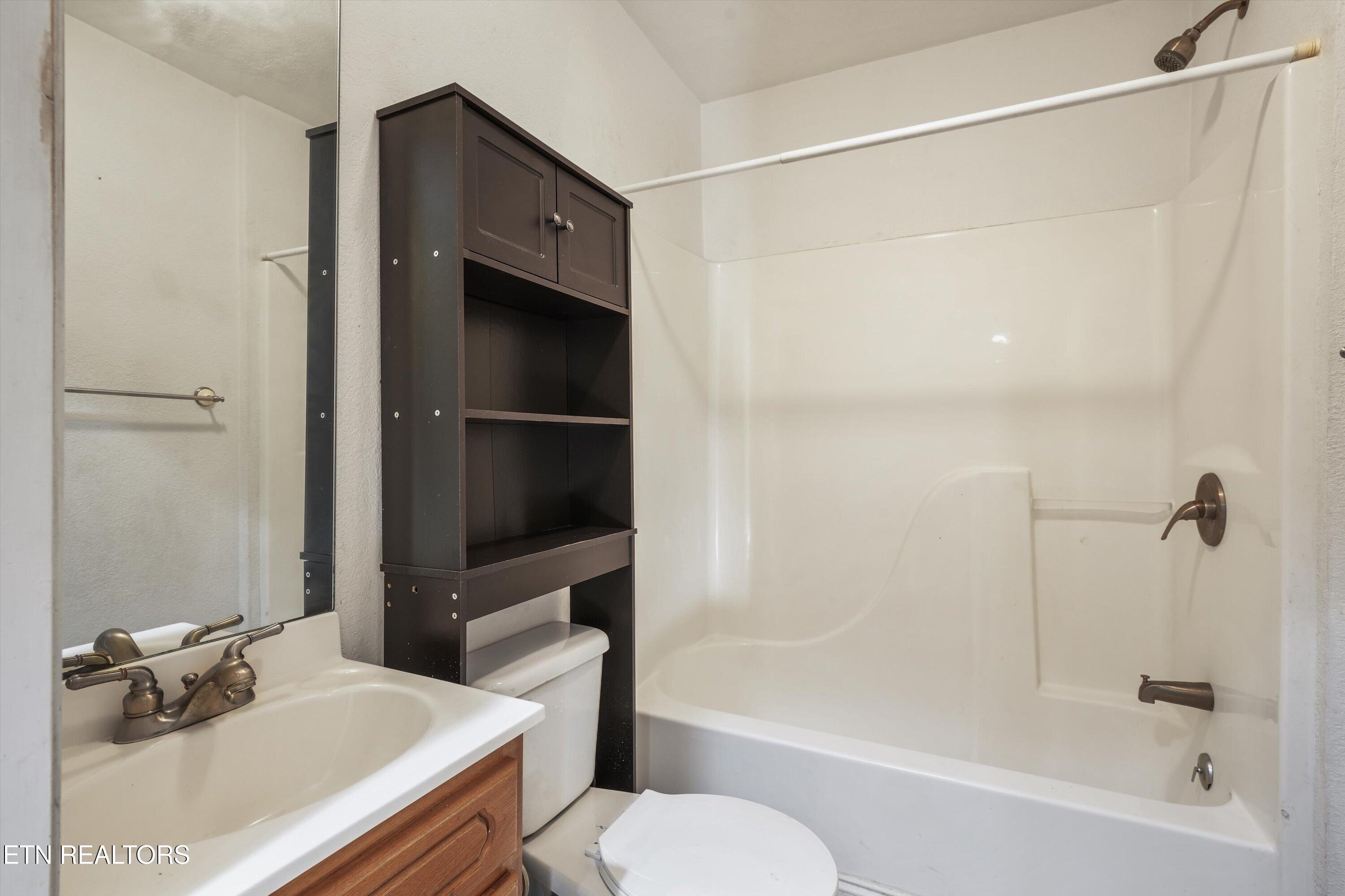 Bathroom with a white tub and shower, dark shelving above the toilet, and a wood-front vanity.