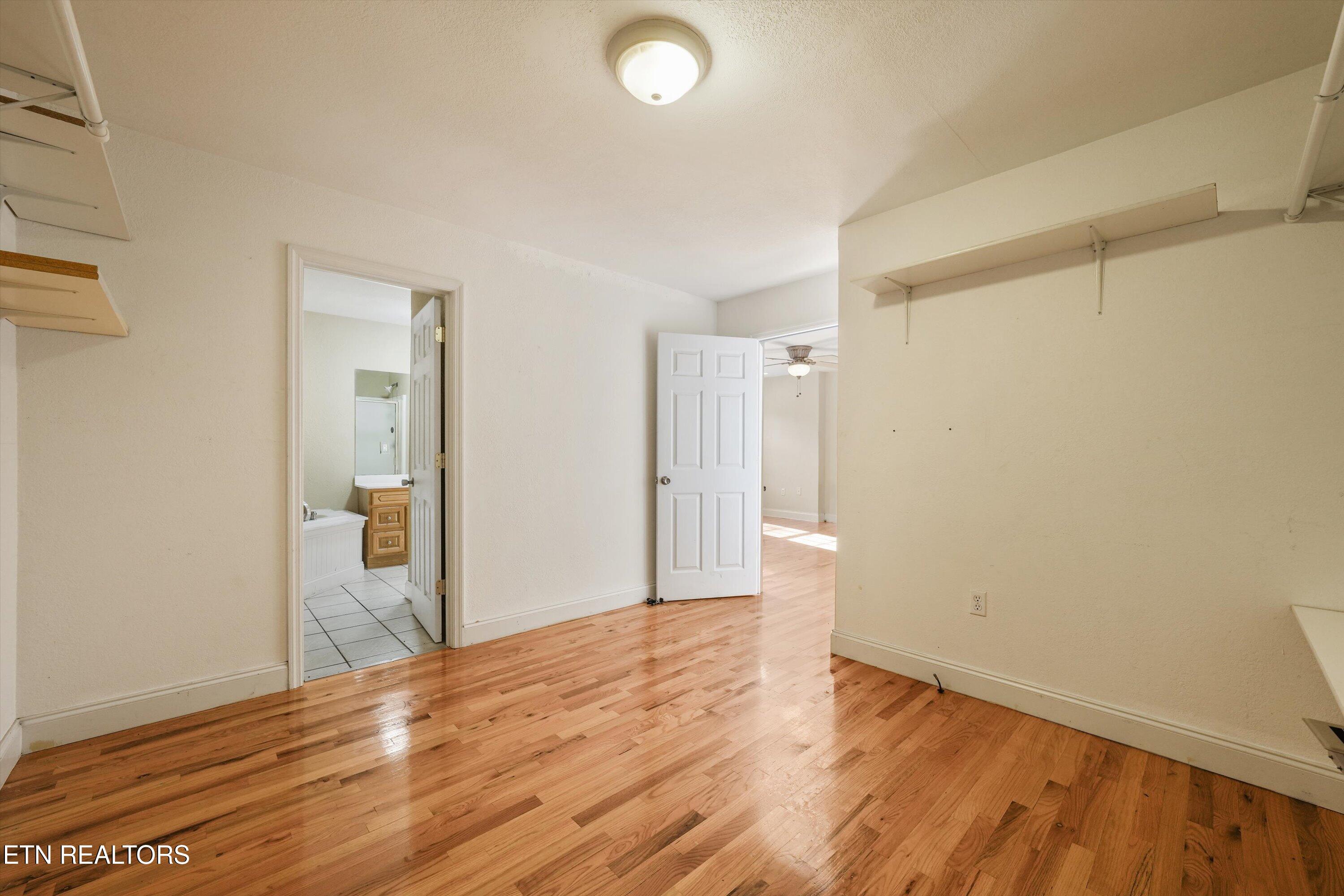 Empty room with hardwood floors and wall shelves, opening to a bathroom and adjoining room.