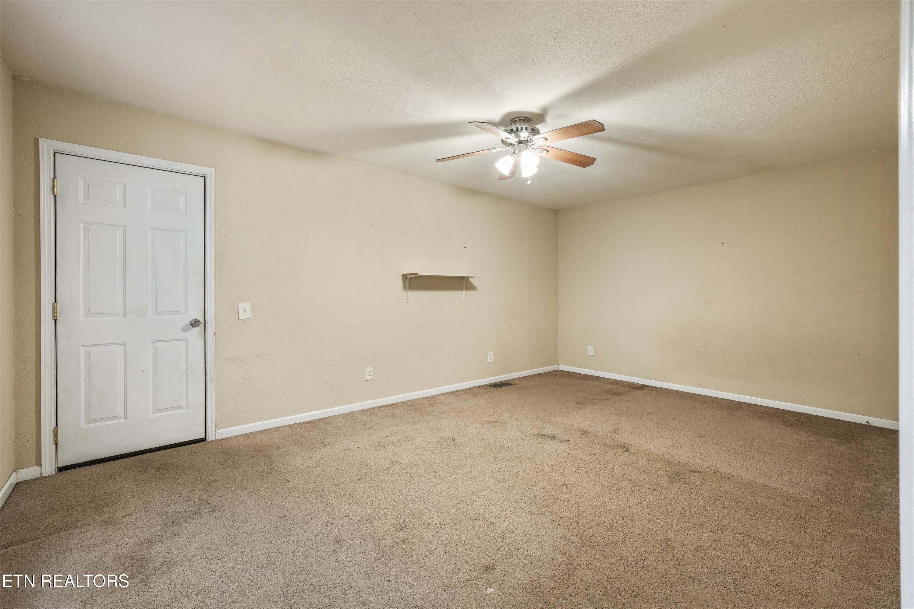 Empty beige room with stained carpet, a white paneled door, and a lit ceiling fan.