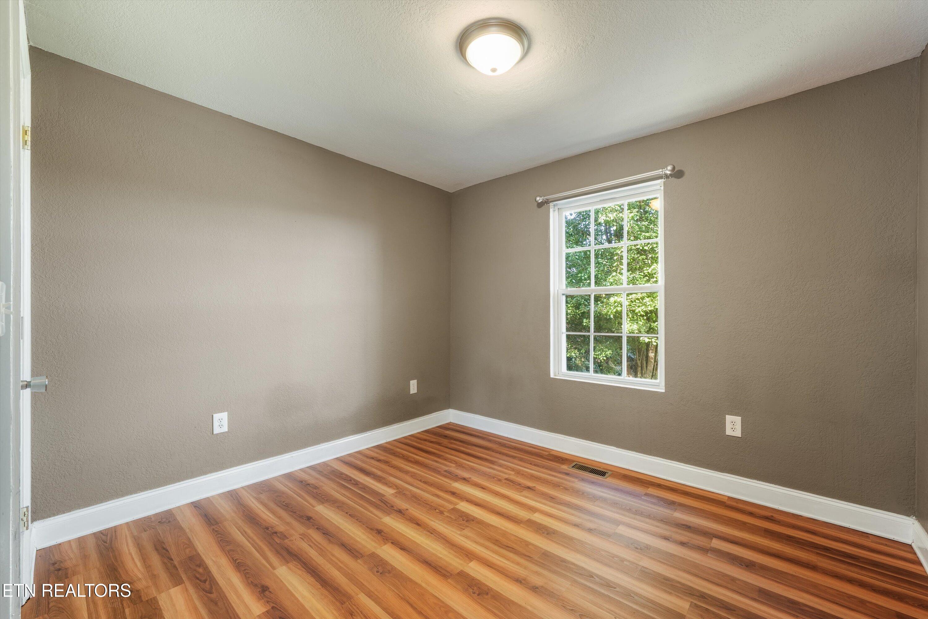 Empty room with warm wood-pattern flooring and a window overlooking leafy trees.