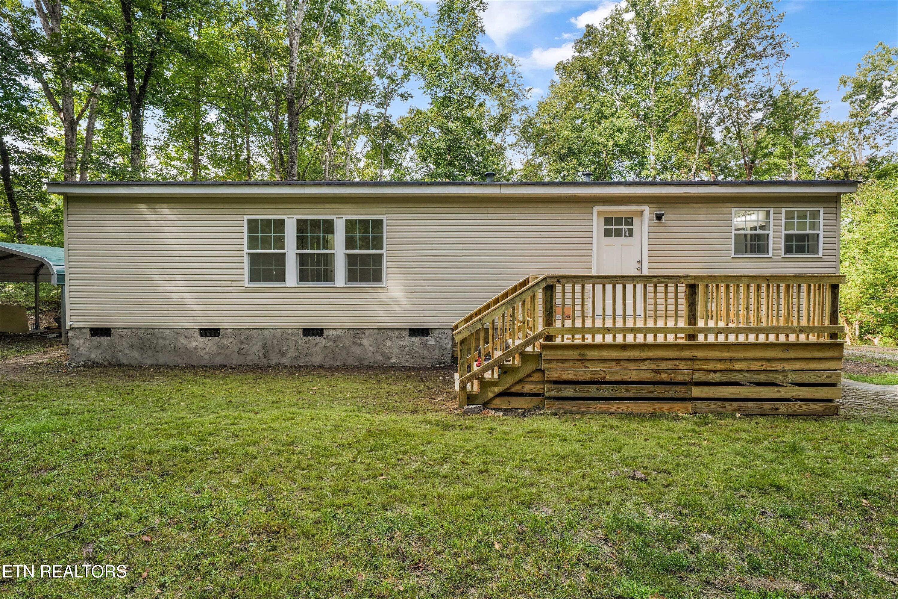 Beige-sided home with a raised wooden deck and stairs, surrounded by a grassy yard and mature trees.