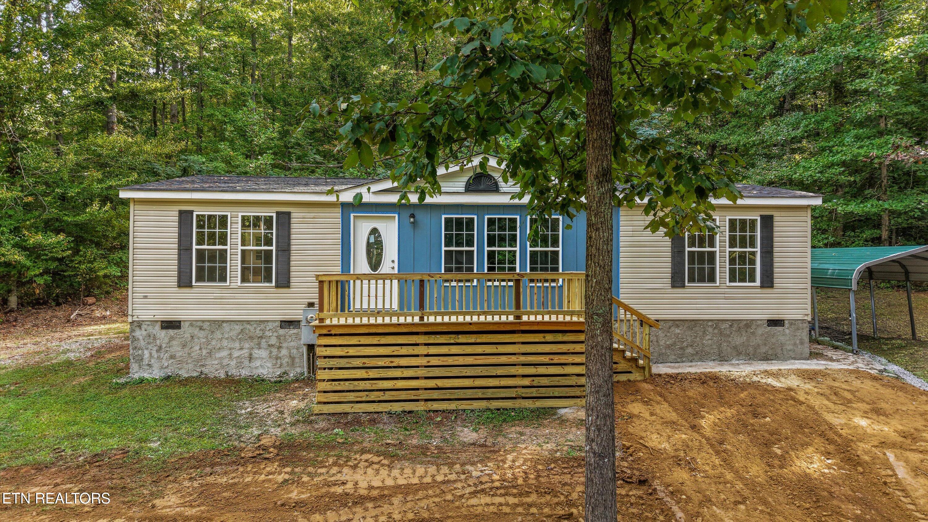 Single-story home with a blue facade, wooden front deck, and carport among trees.