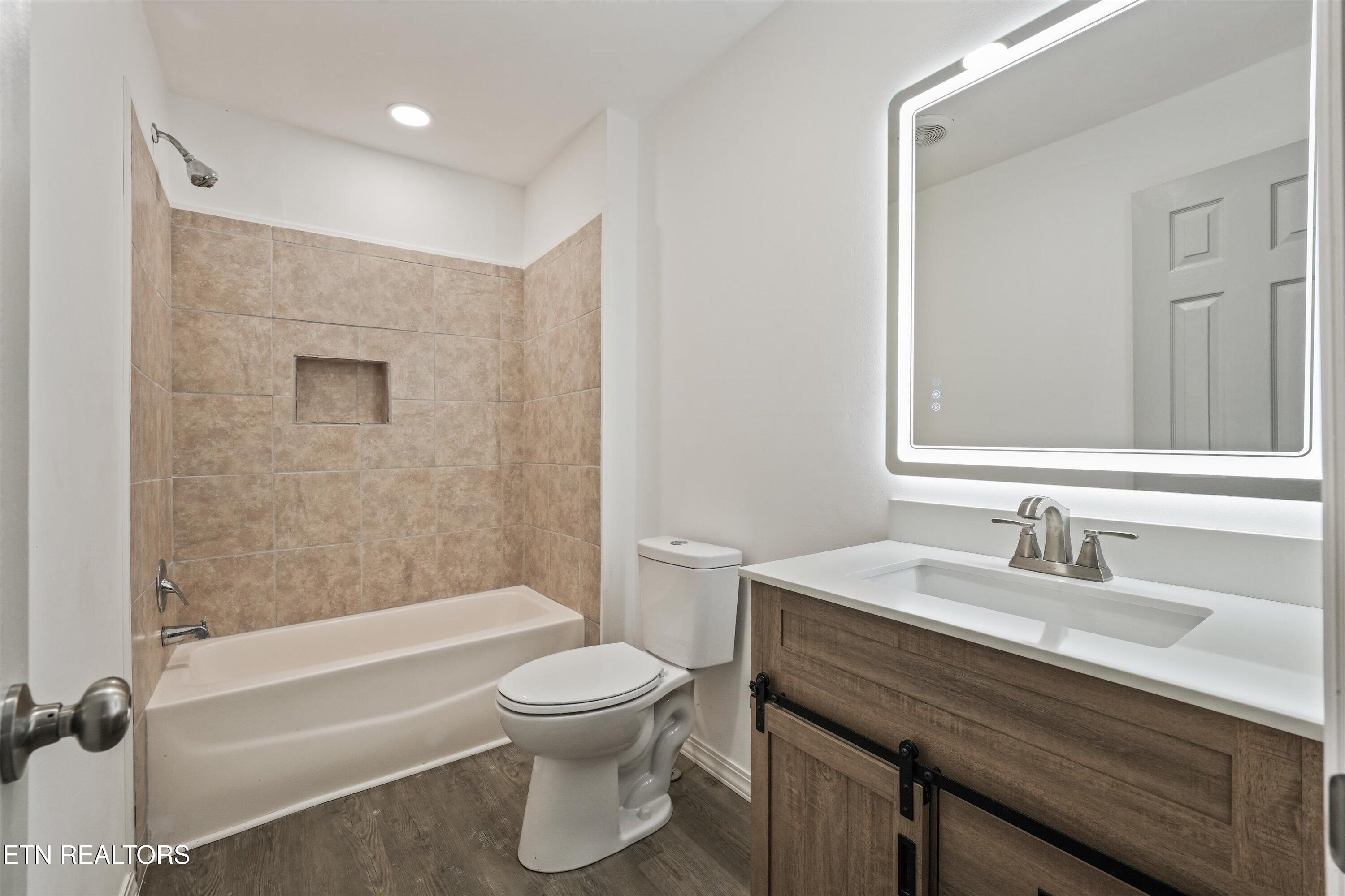Bathroom with a tan-tiled tub and shower, wood-grain vanity, and illuminated mirror.
