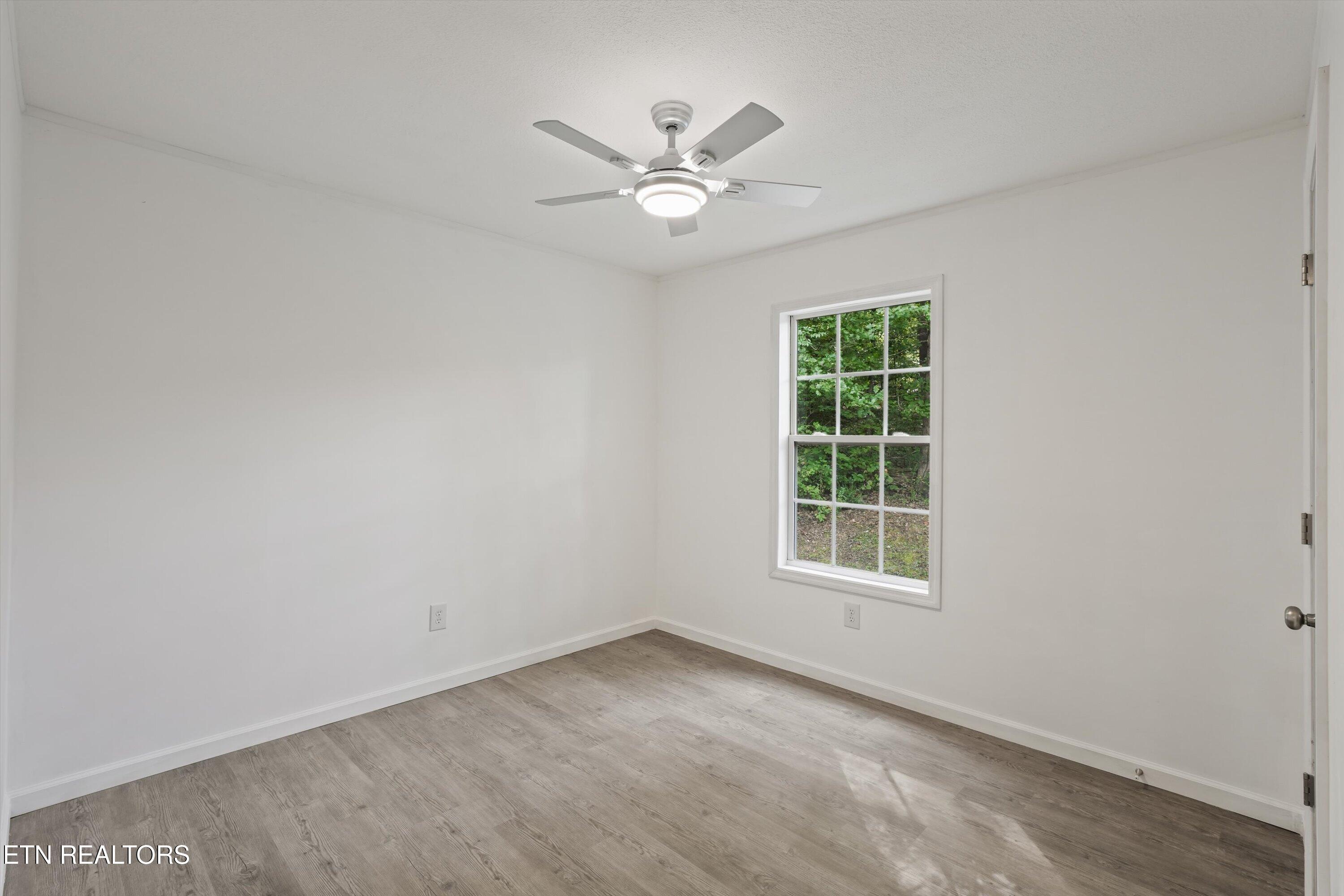 Empty bedroom with gray wood-look flooring, a ceiling fan, and a window facing trees.