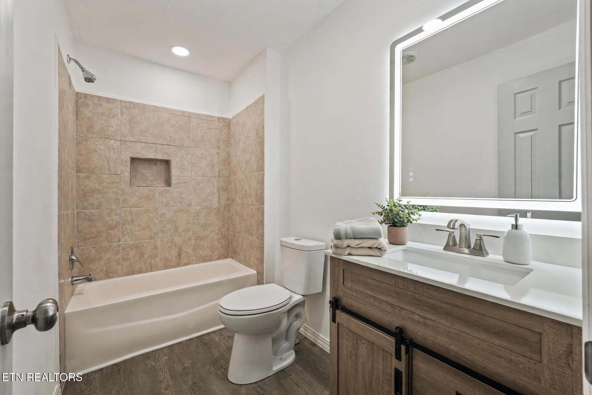 Bathroom with tan tiled tub surround, illuminated mirror, and wood-front vanity.