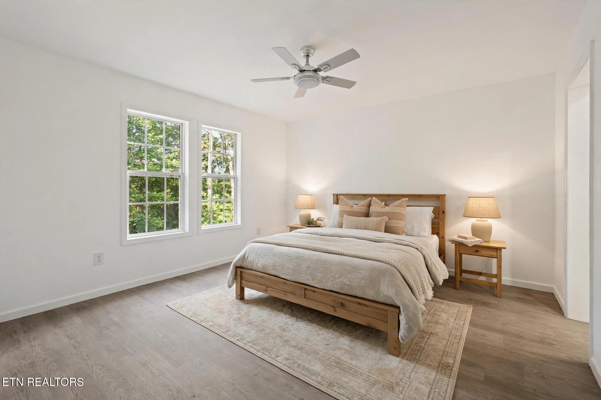 Furnished bedroom with a wood bed, two tree-facing windows, matching bedside tables, and a ceiling fan.