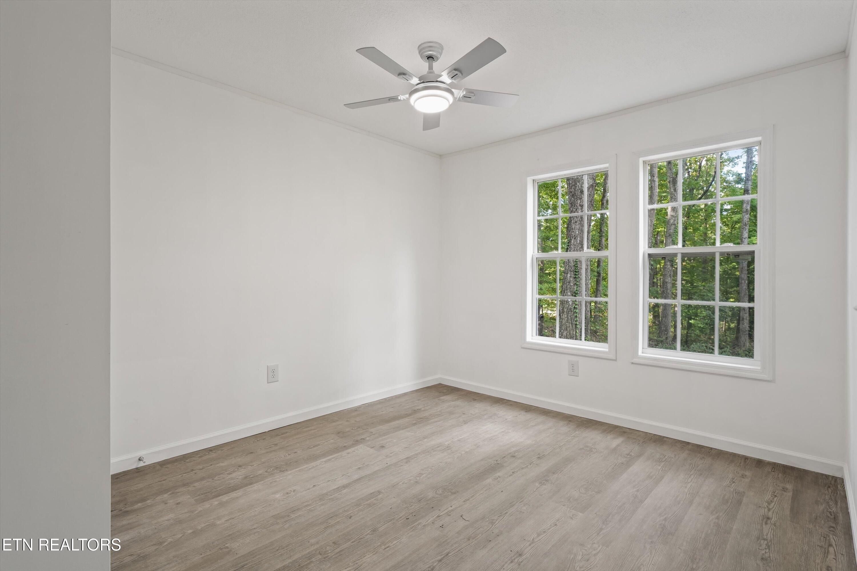 Vacant room with plank flooring, a ceiling fan, and two windows overlooking trees.