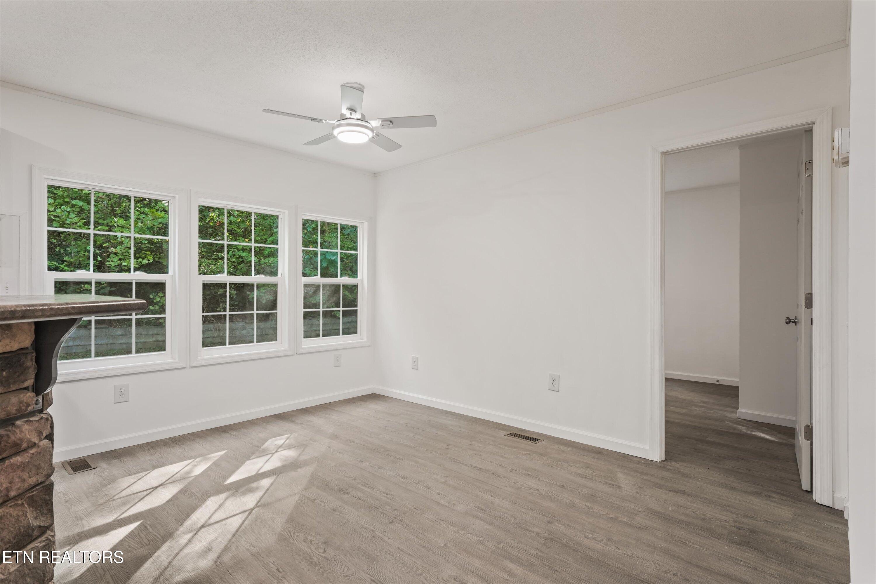Vacant room with three windows, wood-look floors, ceiling fan, and a partial stone fireplace.