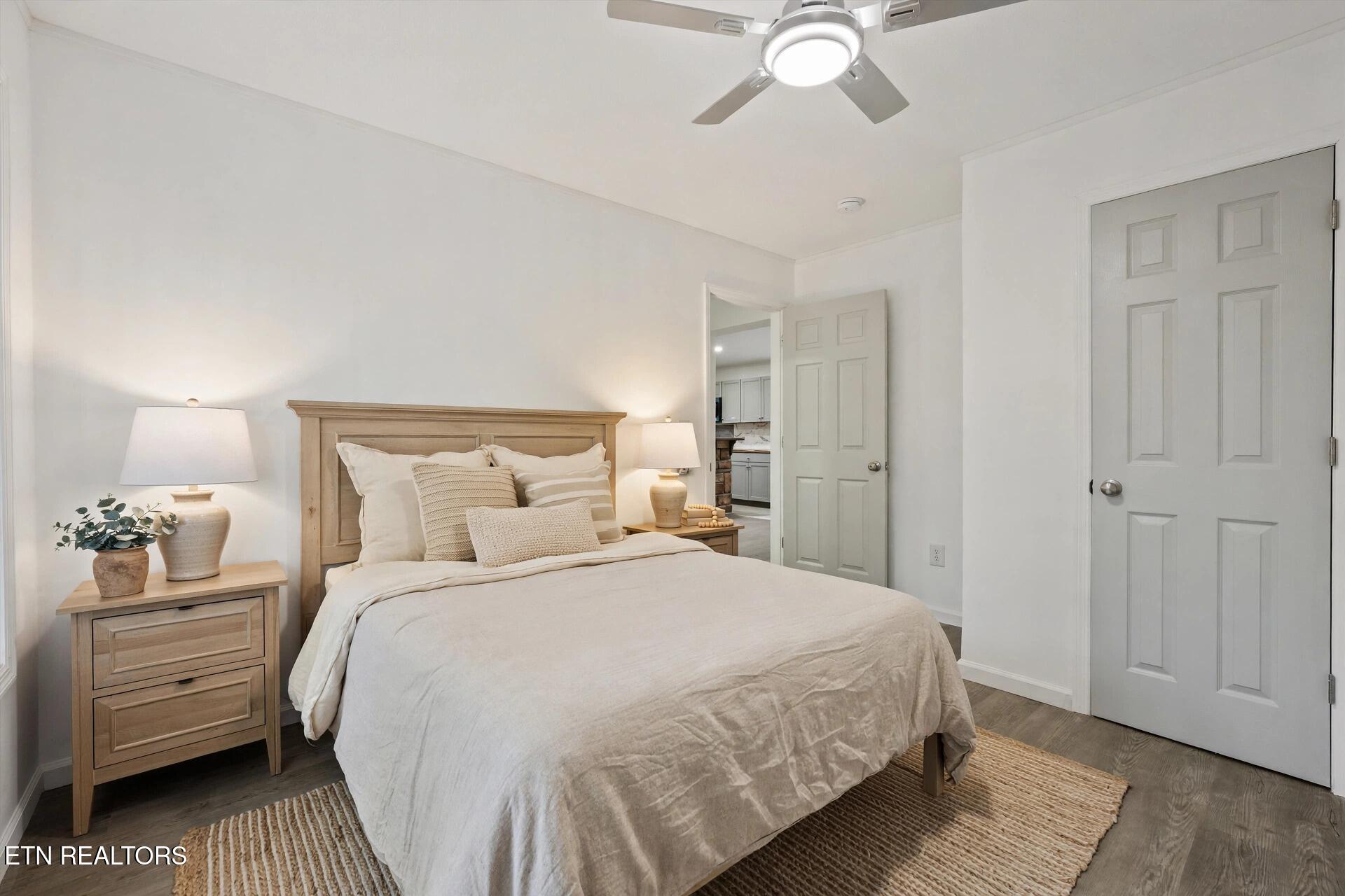Staged bedroom with a light wood bed, cream bedding, woven rugs, and an open doorway to the kitchen.