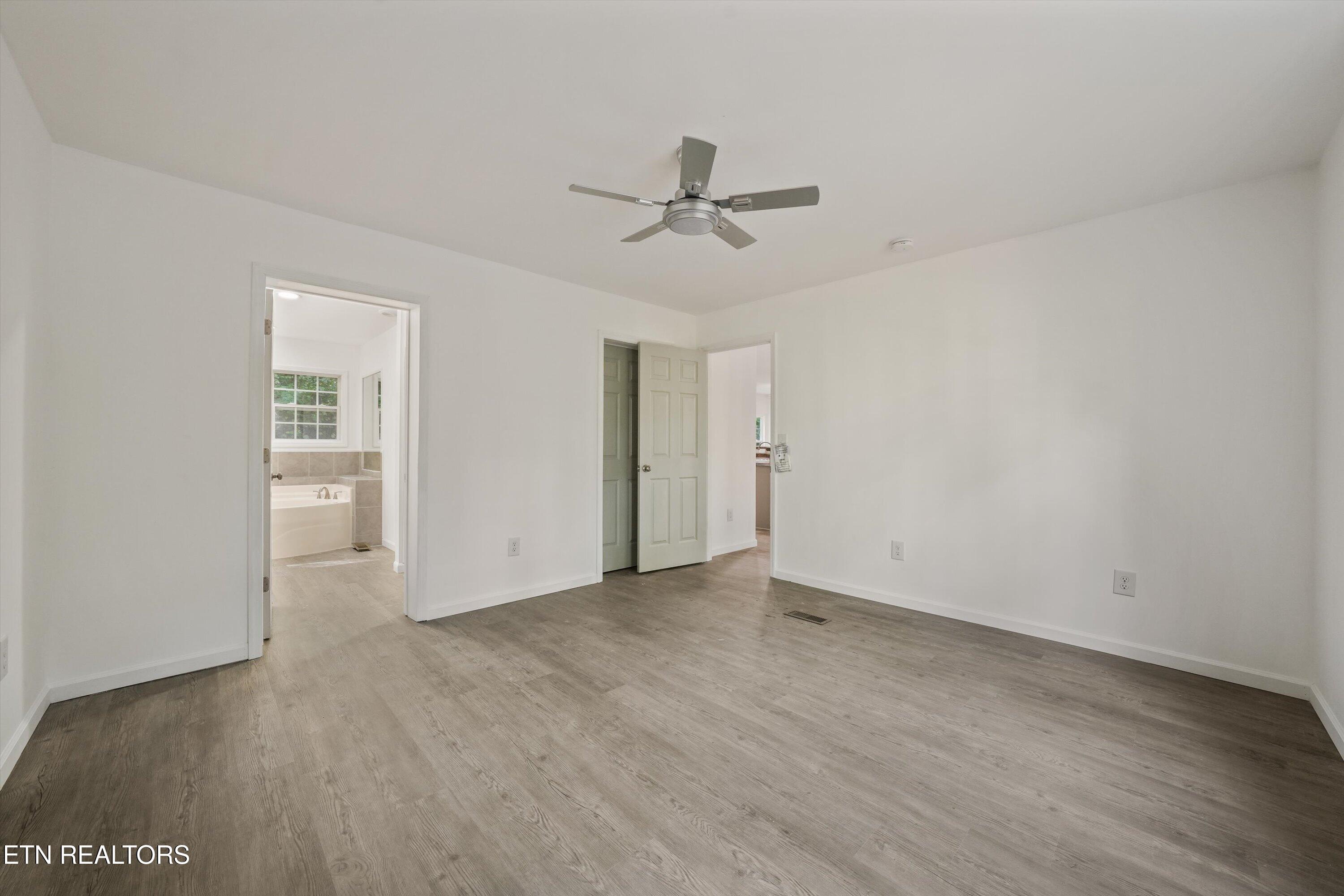 Empty bedroom with gray flooring and a ceiling fan, opening to a bathroom with a tub.
