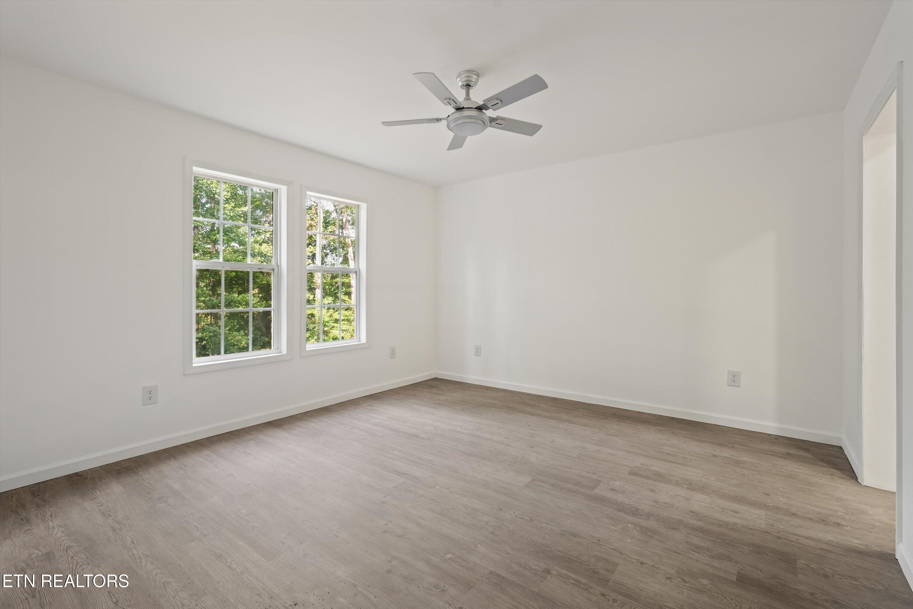 Vacant room with two windows overlooking trees, wood-look flooring, and a ceiling fan.