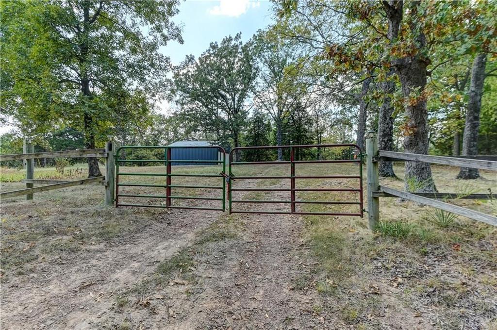 Rural property entrance with double metal gates, a dirt drive, and a blue outbuilding among mature trees.