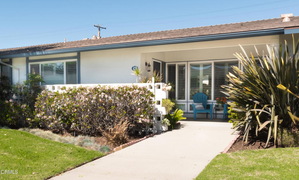 Single-story home with a flowering hedge, landscaped walkway, and blue patio chairs.