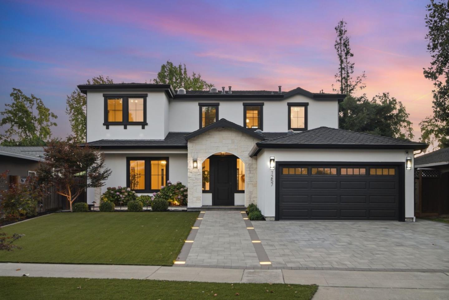 Modern white two-story home with black trim, stone entry, wide garage, and a landscaped front lawn at dusk.