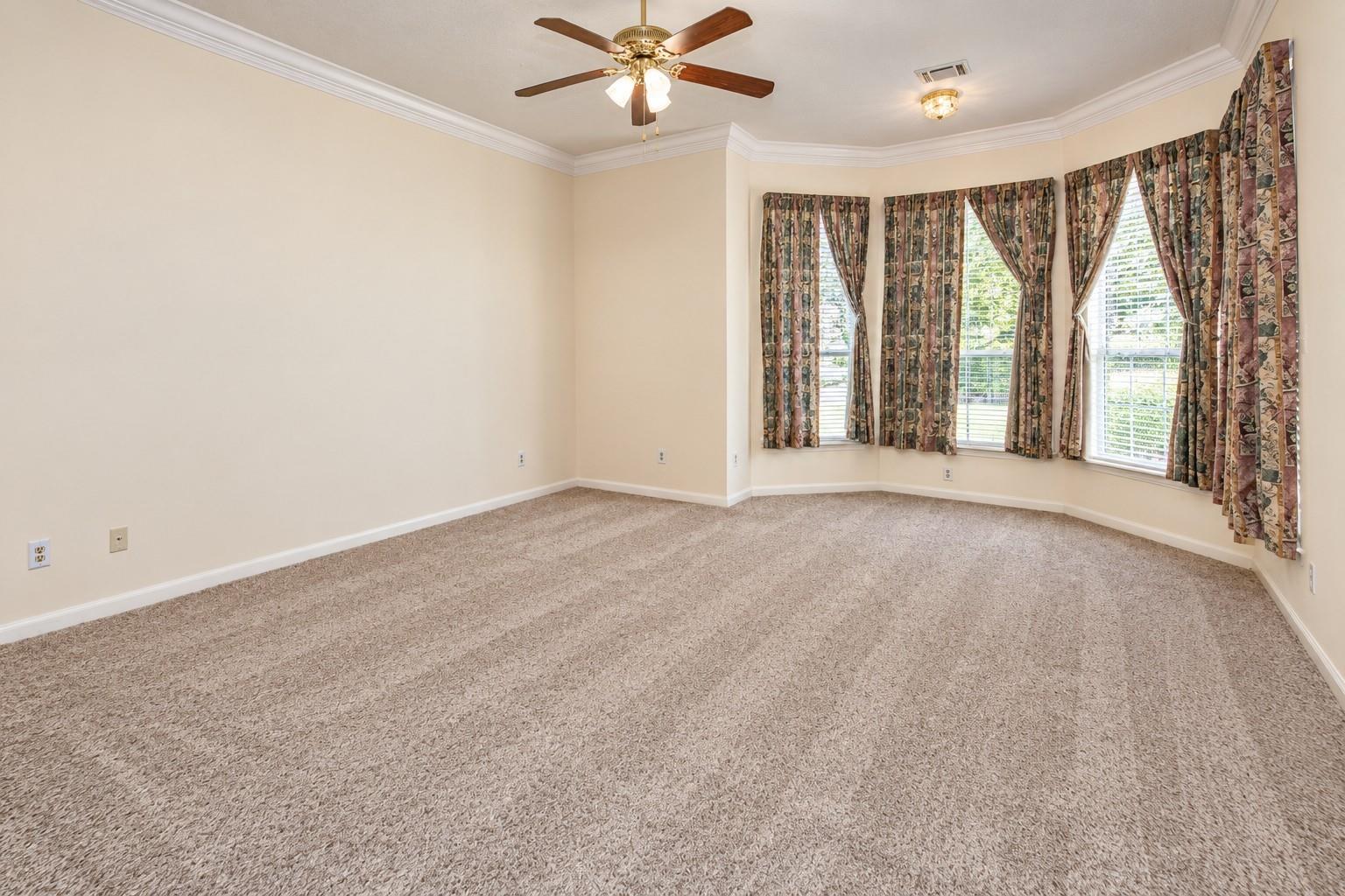 Empty bedroom with beige carpet, a ceiling fan, and curtained bay windows.
