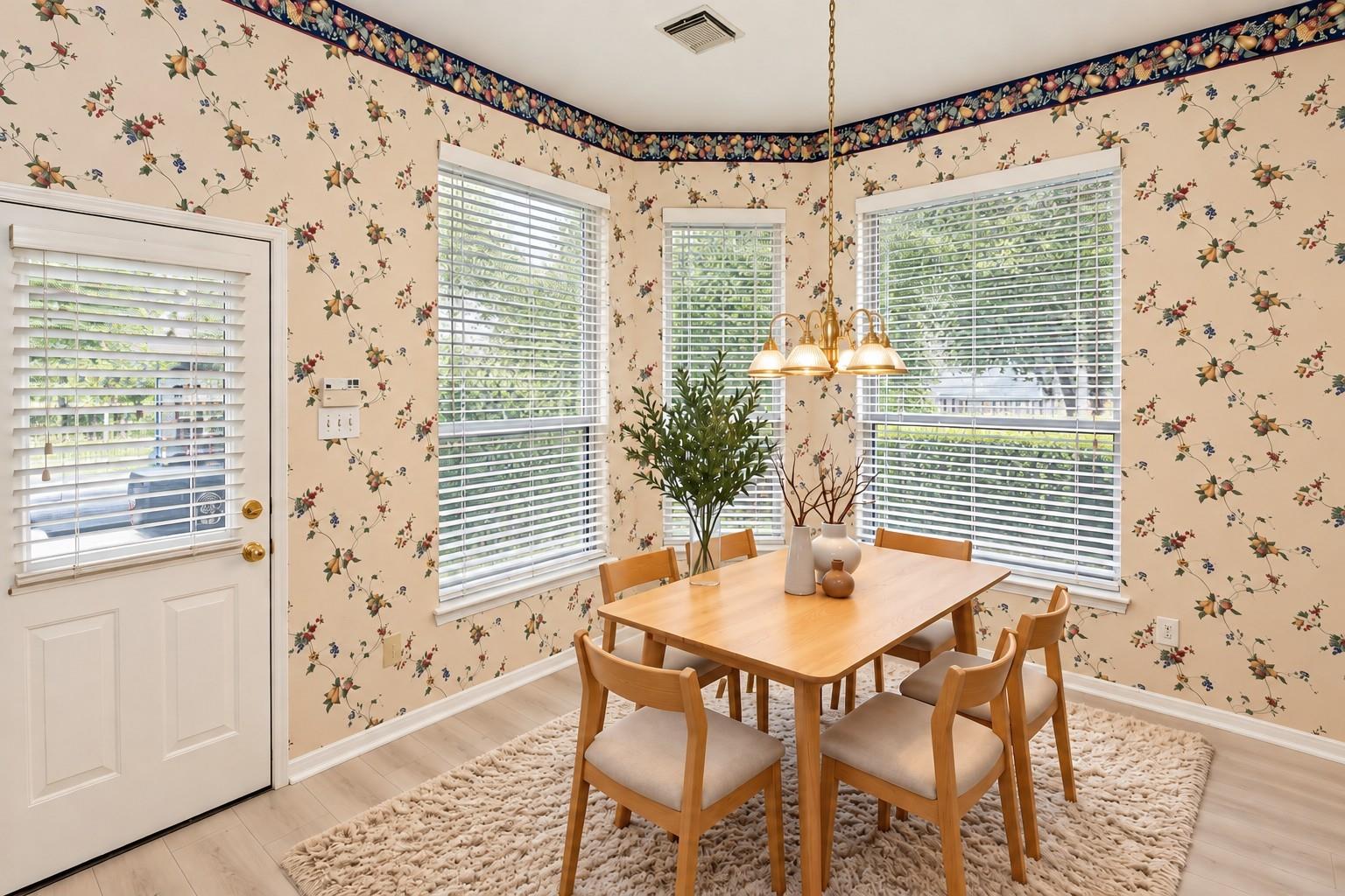 Dining room with floral wallpaper, a wooden table, upholstered chairs, and large windows with white blinds.